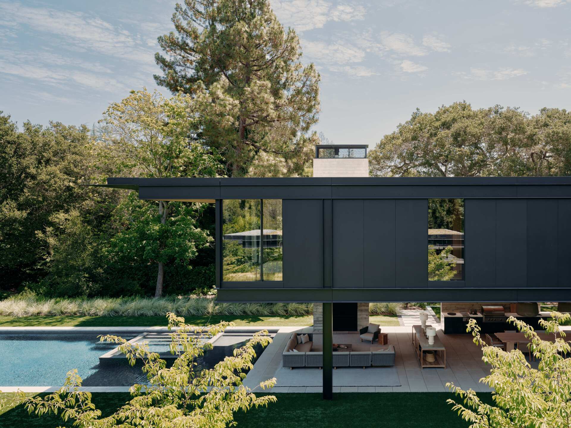 Take a look at this covered outdoor living room at an Olson Kundig home in California. Featuring contemporary outdoor sofas, timber furniture, a fireplace, television, outdoor kitchen, and a beautiful wood-lined ceiling beneath a cantilevered second floor.