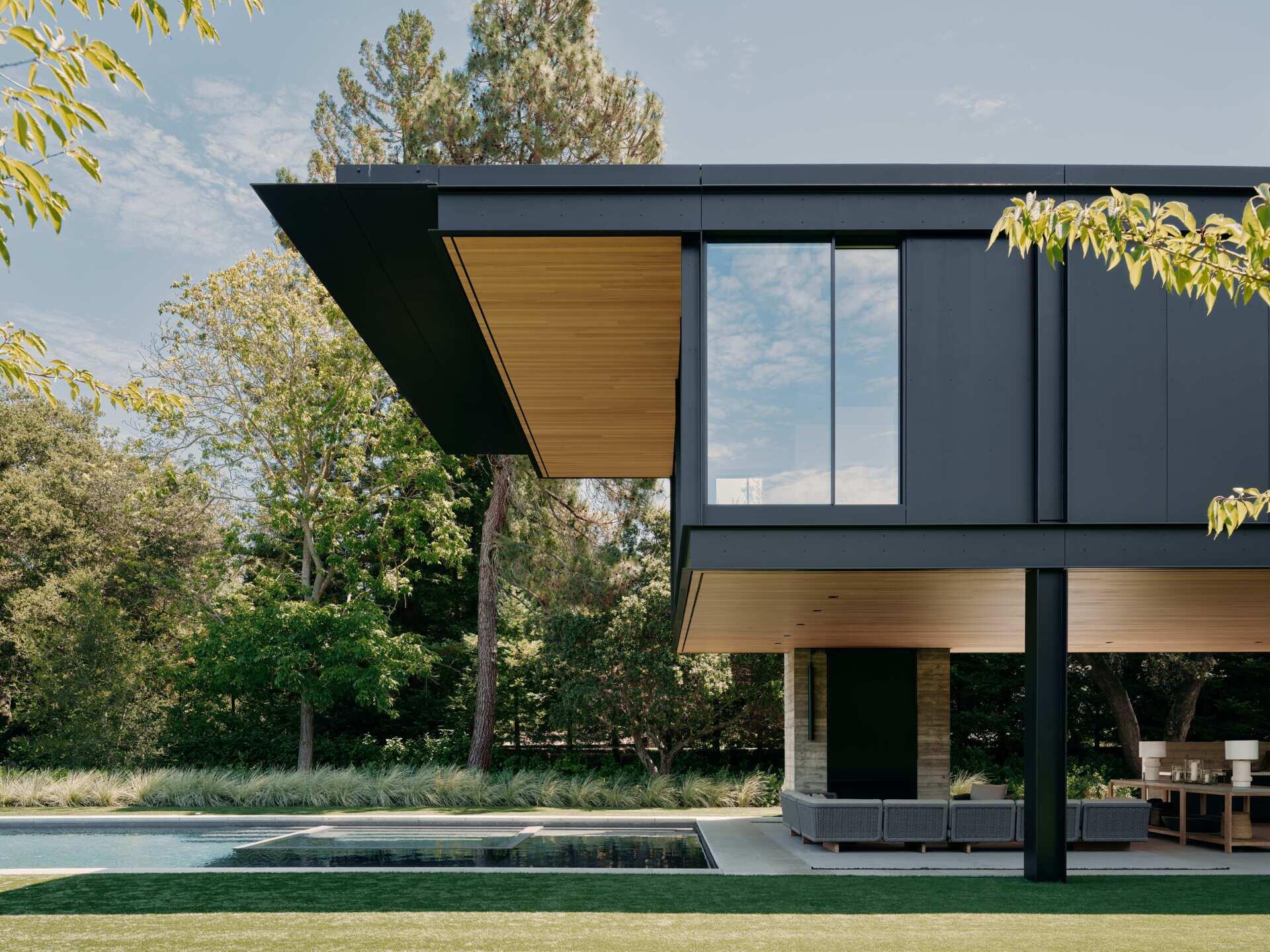Take a look at this covered outdoor living room at an Olson Kundig home in California. Featuring contemporary outdoor sofas, timber furniture, a fireplace, television, outdoor kitchen, and a beautiful wood-lined ceiling beneath a cantilevered second floor.