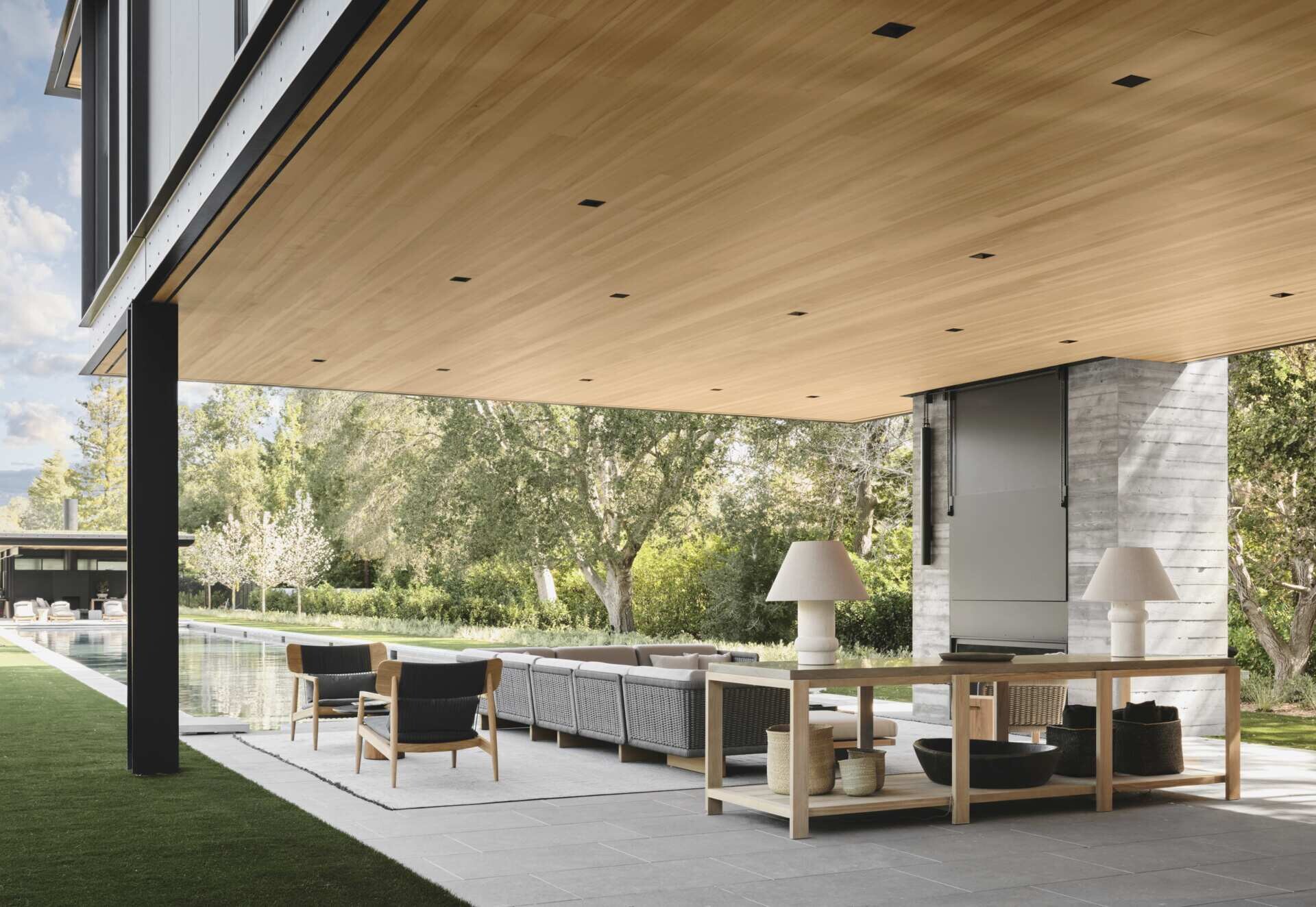 Take a look at this covered outdoor living room at an Olson Kundig home in California. Featuring contemporary outdoor sofas, timber furniture, a fireplace, television, outdoor kitchen, and a beautiful wood-lined ceiling beneath a cantilevered second floor.