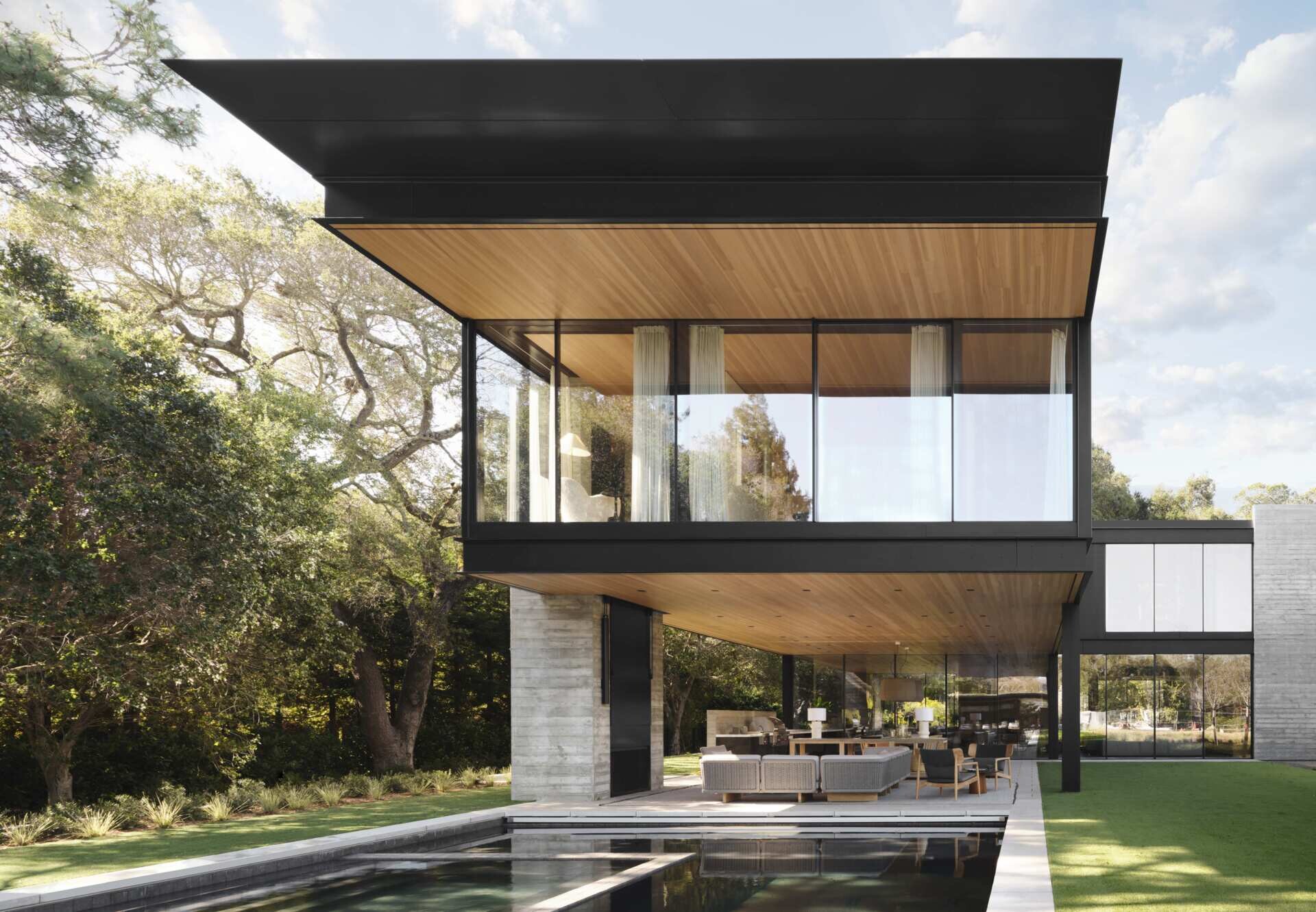 Take a look at this covered outdoor living room at an Olson Kundig home in California. Featuring contemporary outdoor sofas, timber furniture, a fireplace, television, outdoor kitchen, and a beautiful wood-lined ceiling beneath a cantilevered second floor.