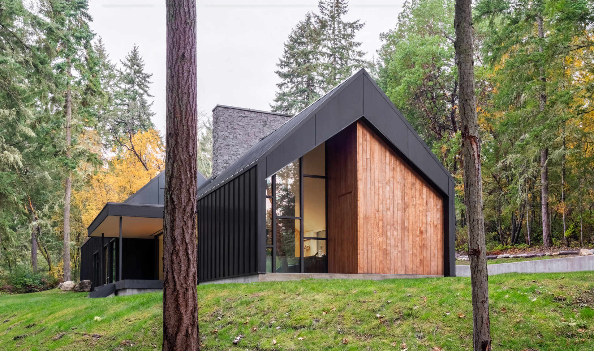 This modern gabled house on Vashon Island features black standing-seam metal, recessed timber walls, floor-to-ceiling glass, and a narrow footprint designed to preserve the surrounding forest.