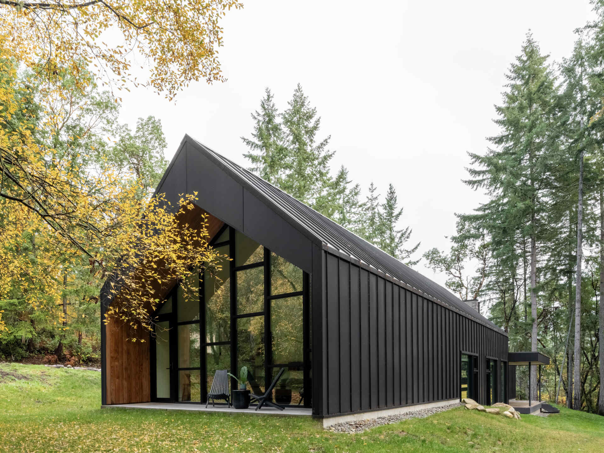 This modern gabled house on Vashon Island features black standing-seam metal, recessed timber walls, floor-to-ceiling glass, and a narrow footprint designed to preserve the surrounding forest.