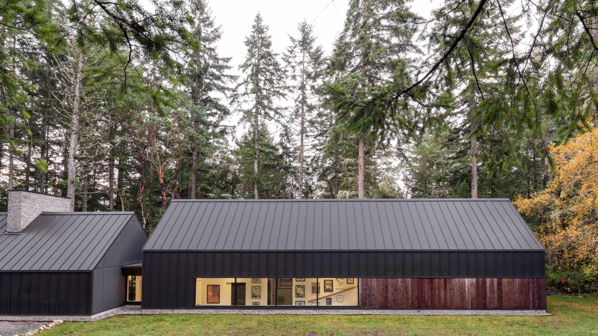 This modern gabled house on Vashon Island features black standing-seam metal, recessed timber walls, floor-to-ceiling glass, and a narrow footprint designed to preserve the surrounding forest.