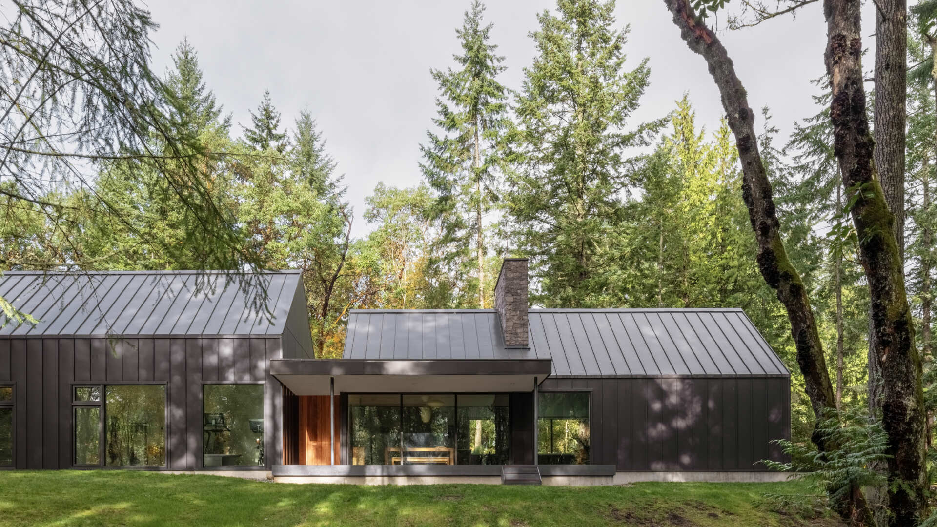 A covered porch extends from this modern forest house, featuring sliding glass doors, a minimalist roof canopy, and views of the surrounding trees on Vashon Island.