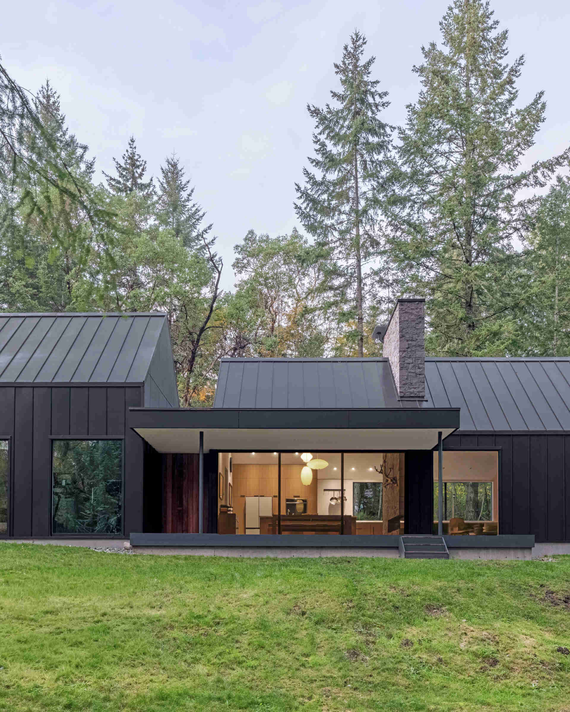 A covered porch extends from this modern forest house, featuring sliding glass doors, a minimalist roof canopy, and views of the surrounding trees on Vashon Island.