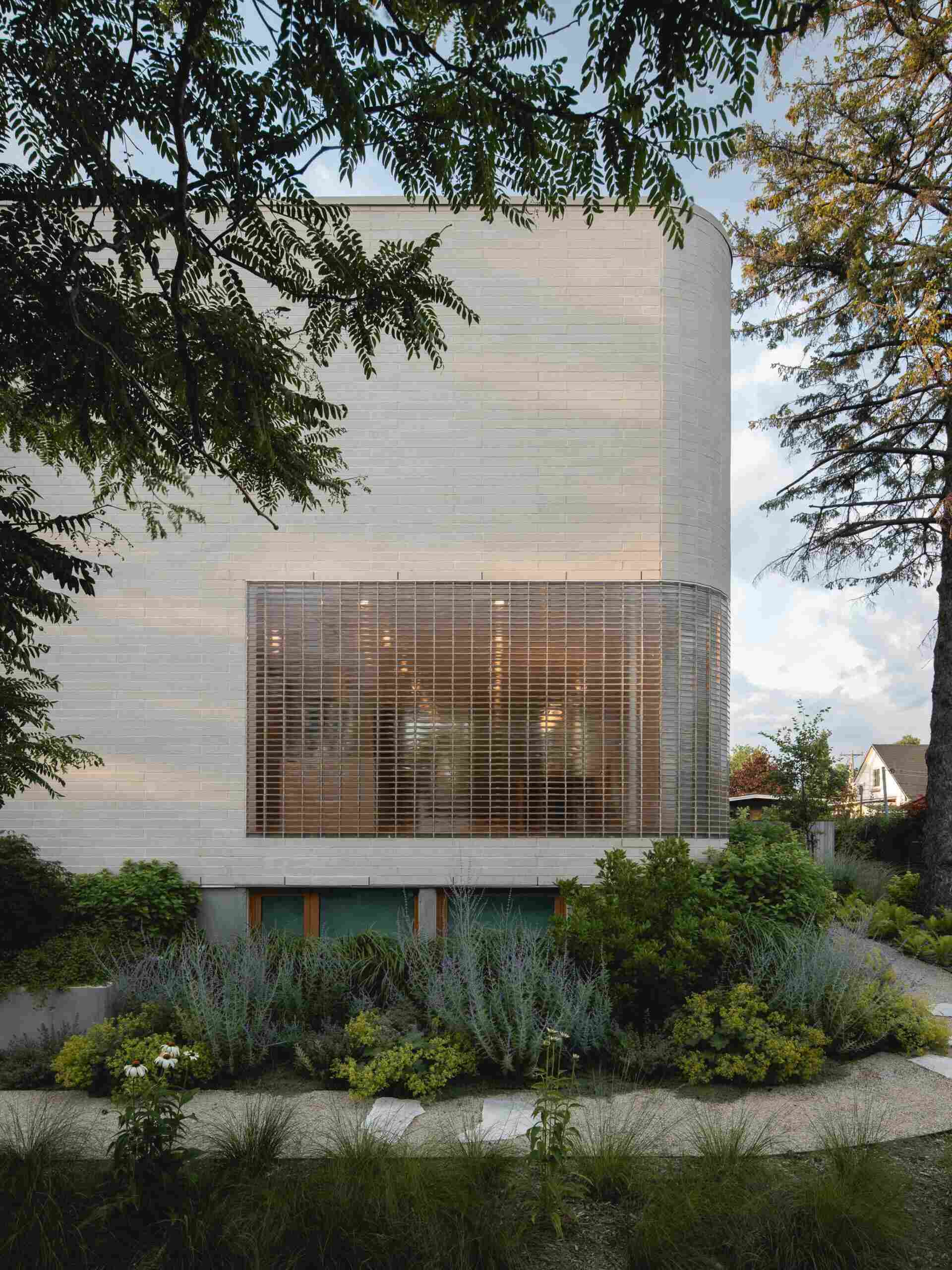 Explore the contemporary façade of The Lens in Montreal, designed by MU Architecture. Pale beige brick, rounded architectural details, a curved glass-brick wall, and lush landscaping create a private modern family home.
