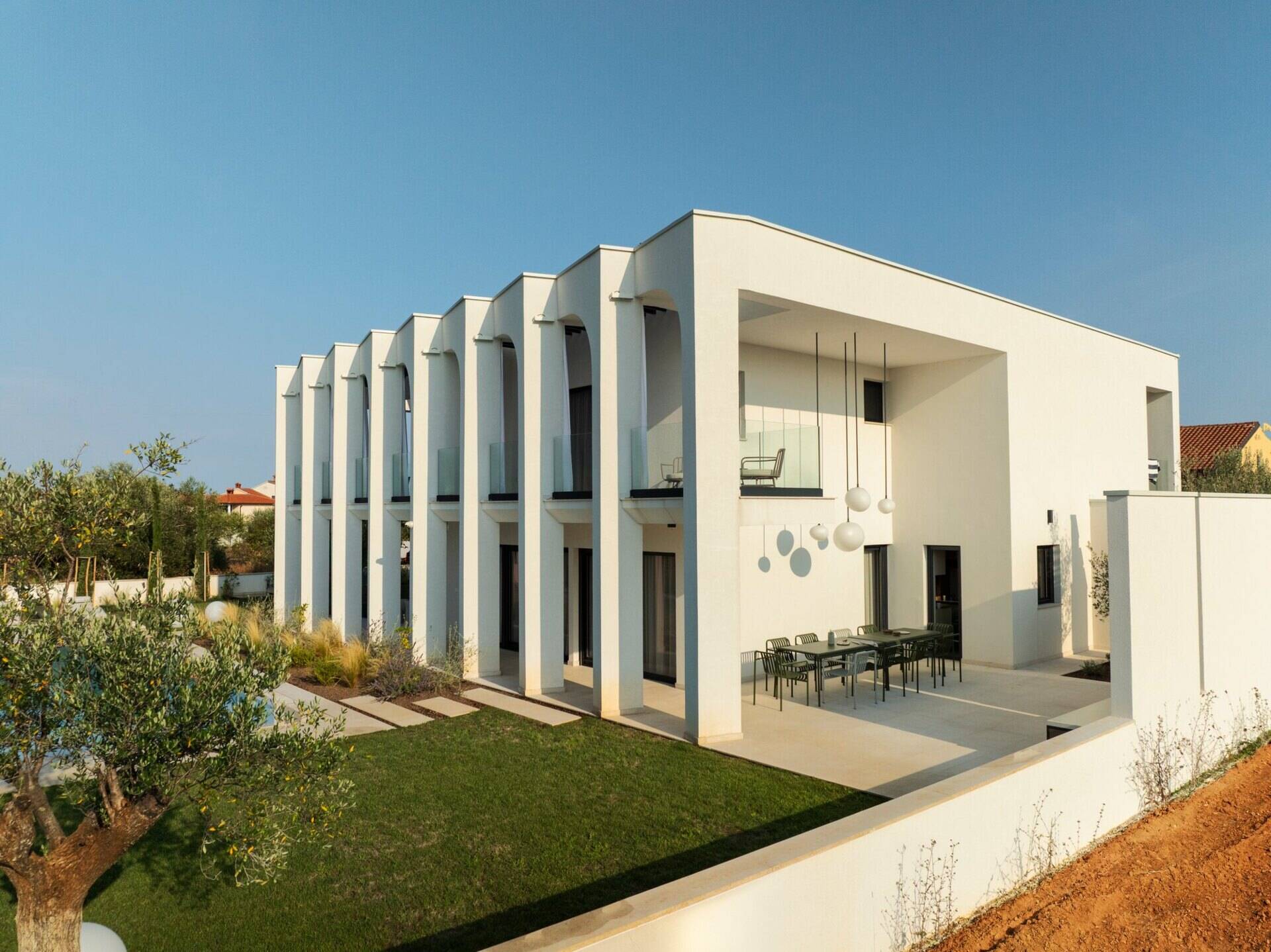 Take a look at the outdoor patios of Arc House in Croatia, featuring a Mediterranean dining terrace with an outdoor kitchen, modern green furniture, globe pendant lights and a covered lounge with white seating and garden views.