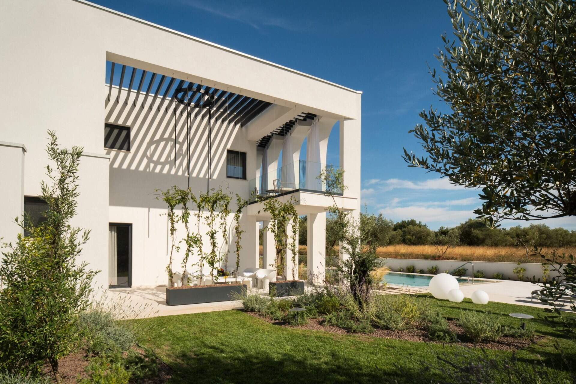 Take a look at the outdoor patios of Arc House in Croatia, featuring a Mediterranean dining terrace with an outdoor kitchen, modern green furniture, globe pendant lights and a covered lounge with white seating and garden views.