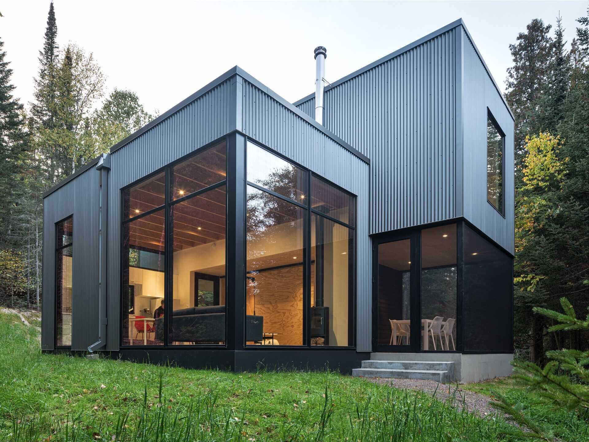 A modern lakeside cabin with corrugated metal siding, floor-to-ceiling corner windows, and a screened porch beneath the upper floor. Warm interior lighting contrasts with the surrounding forest.