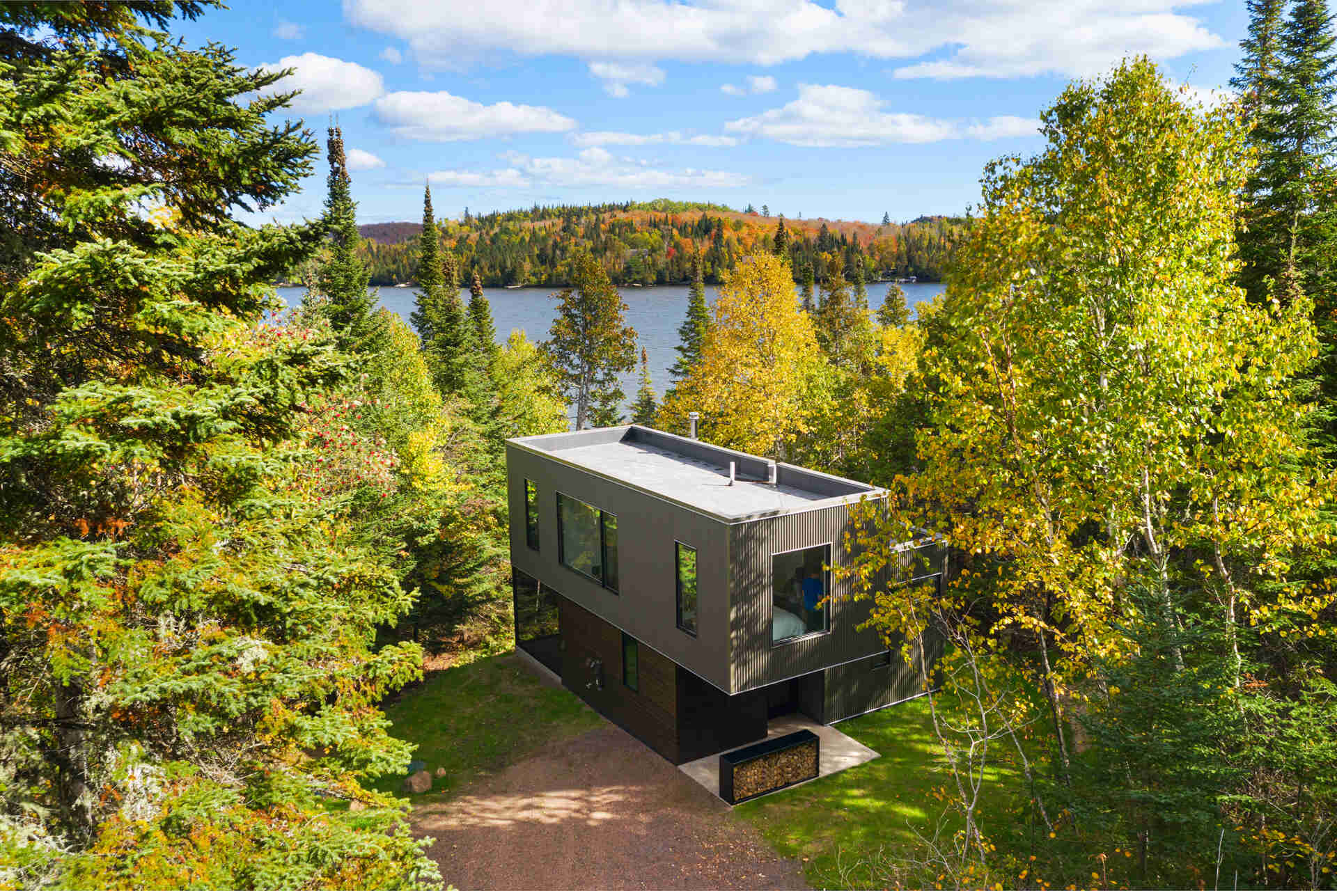 An aerial view of a small modern cabin nestled among autumn trees beside Caribou Lake in Minnesota. The two-story building has a flat roof, dark metal exterior, and large windows overlooking the wooded property.