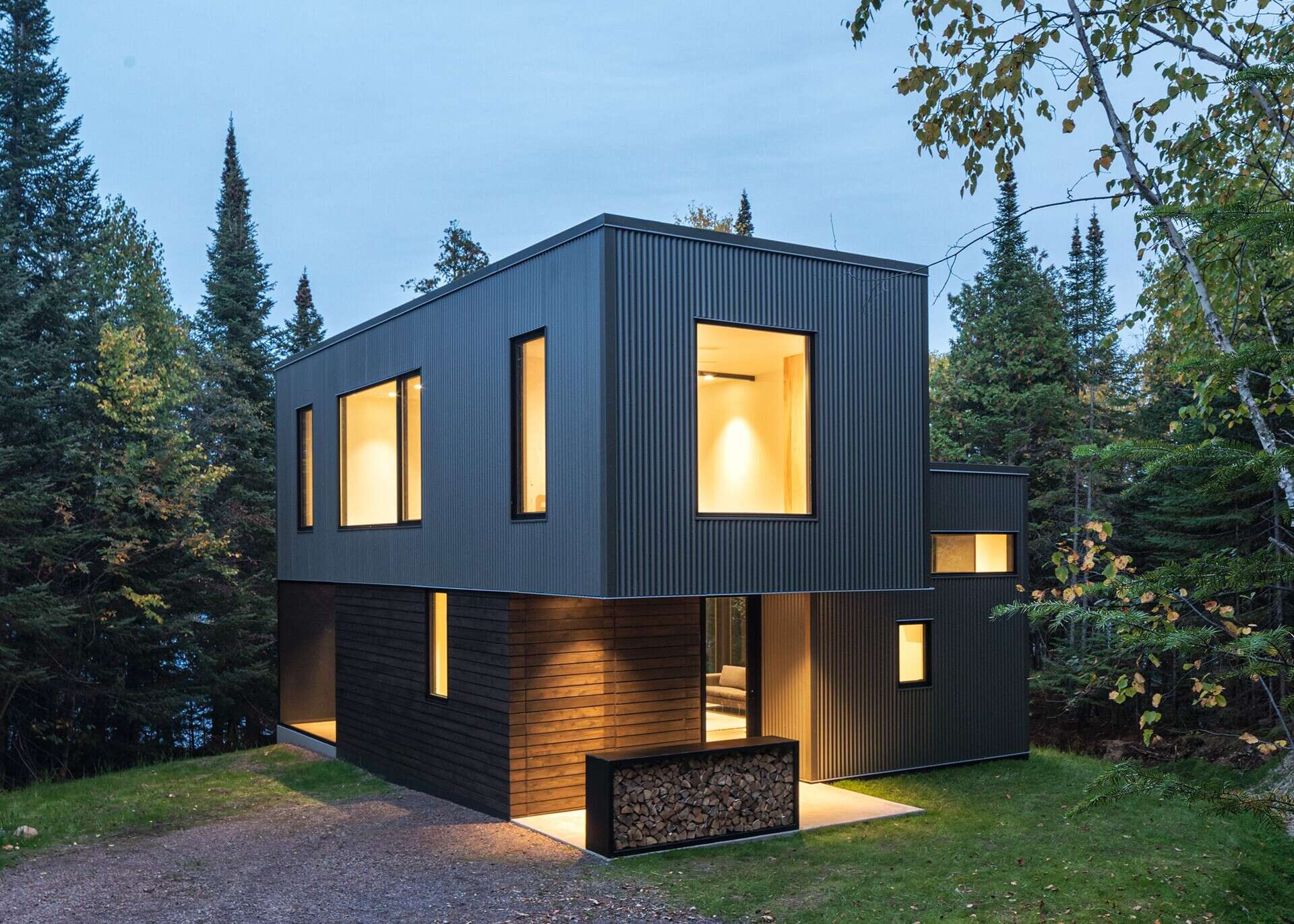A modern cabin exterior featuring a cantilevered second floor, dark corrugated metal siding, and a sheltered entrance. A rectangular steel firewood storage box sits beside the doorway.