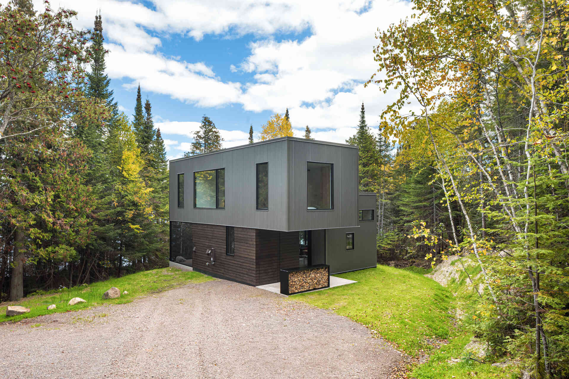 A compact two-story cabin with corrugated metal siding, dark cedar cladding, and a cantilevered upper floor. A gravel driveway leads to the sheltered entrance, surrounded by trees and natural landscaping.