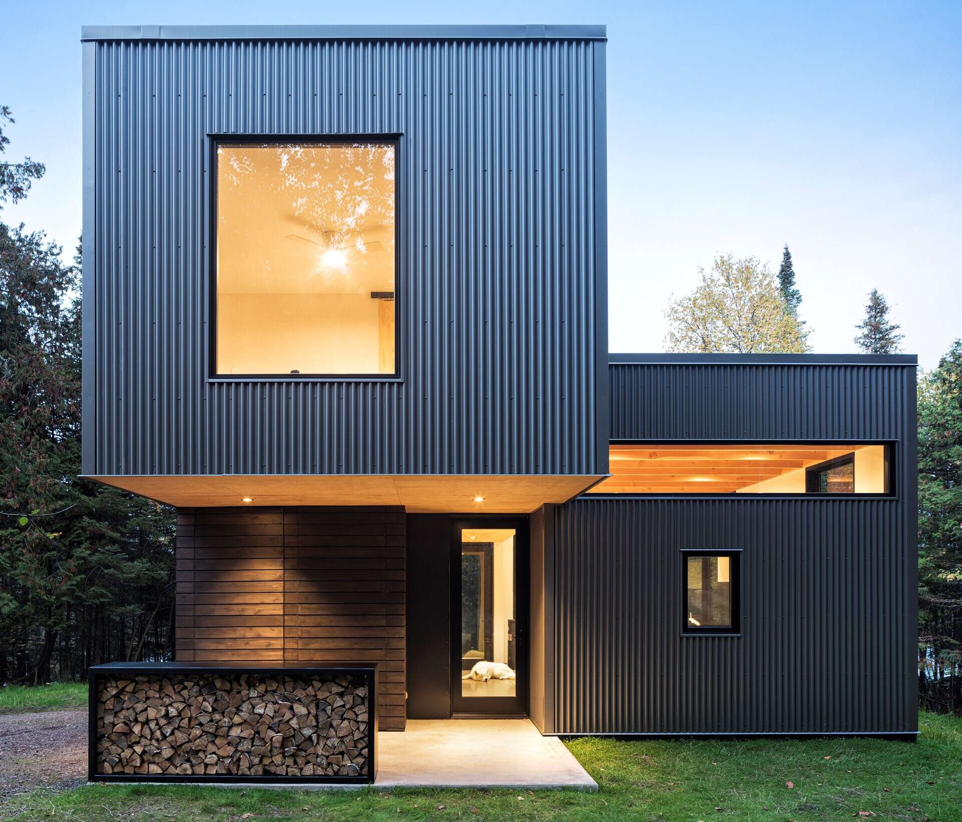 A built-in steel firewood storage box filled with neatly stacked logs beside the entrance of a modern cabin. Dark horizontal cedar siding and a glass entrance door complete the exterior detail.