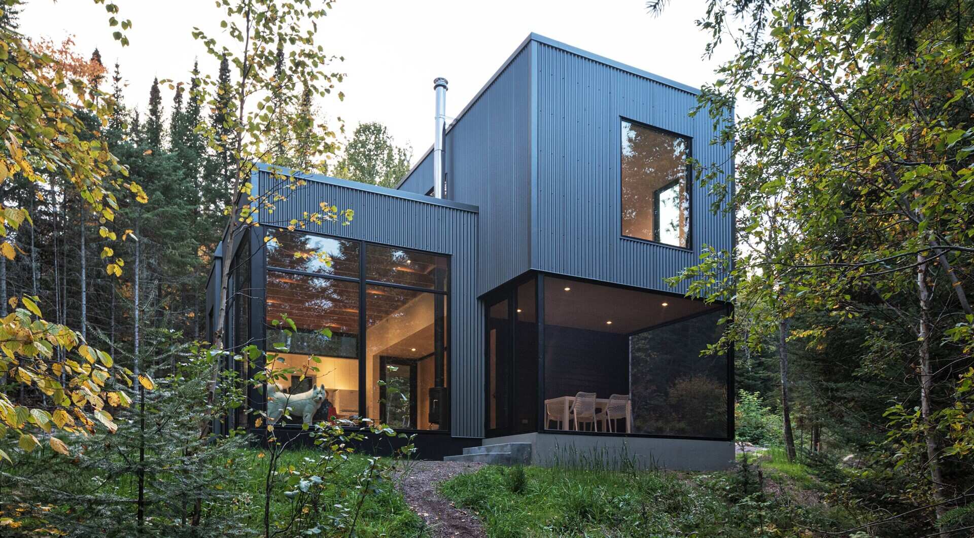 A modern cabin surrounded by trees, featuring dark corrugated metal siding, large corner windows, and a screened dining porch. The projecting upper floor creates a covered outdoor area below.
