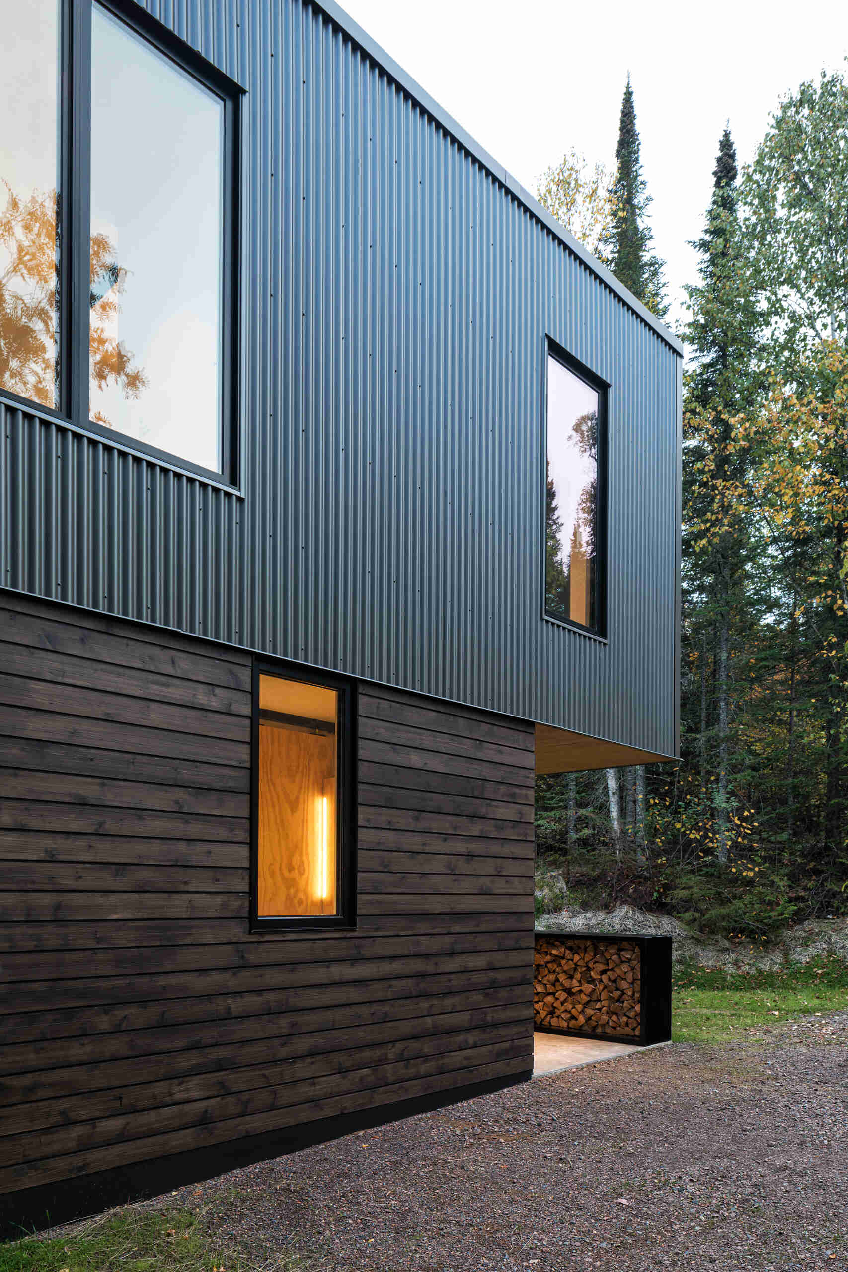 A side view of a contemporary cabin featuring corrugated metal siding above dark-stained cedar cladding. Tall rectangular windows and an overhanging upper floor define the exterior.