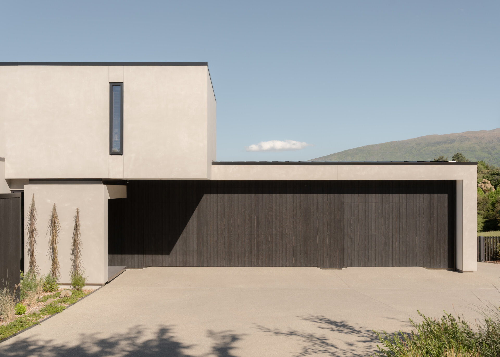 A contemporary house in W?naka, New Zealand, designed by Condon Scott Architects, featuring a cantilevered upper level, pale plaster exterior, black wood cladding, and modern geometric architecture.