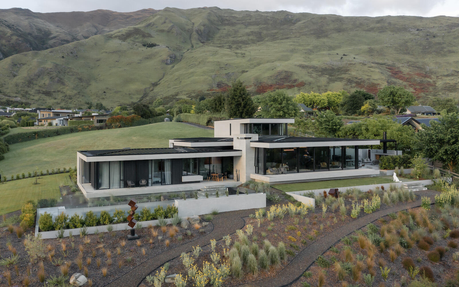 Explore a contemporary hillside house in W?naka featuring stepped architecture, floating concrete terraces, floor-to-ceiling windows, mountain views, and landscaped gardens with sculptural artworks.