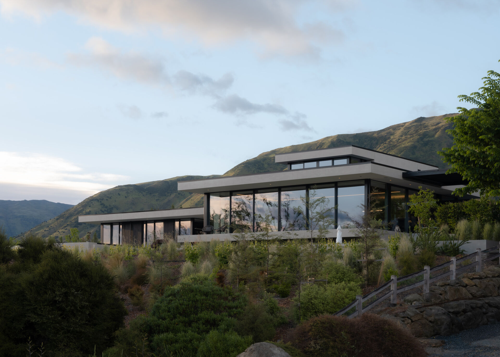 Explore a contemporary hillside house in W?naka featuring stepped architecture, floating concrete terraces, floor-to-ceiling windows, mountain views, and landscaped gardens with sculptural artworks.