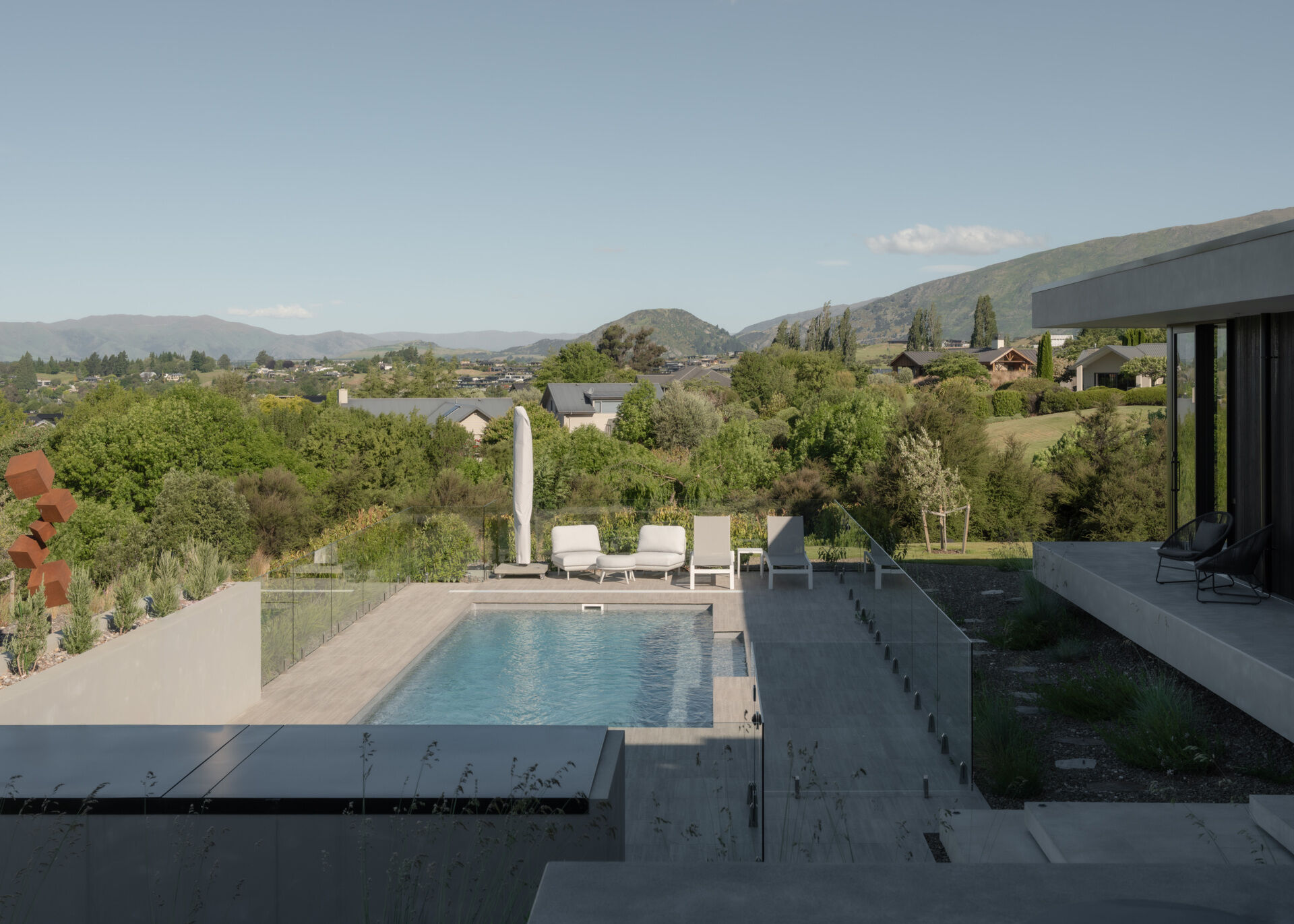 A modern outdoor swimming pool in W?naka, New Zealand, with pale stone-colored paving, frameless glass fencing, white sun loungers, and expansive mountain views.