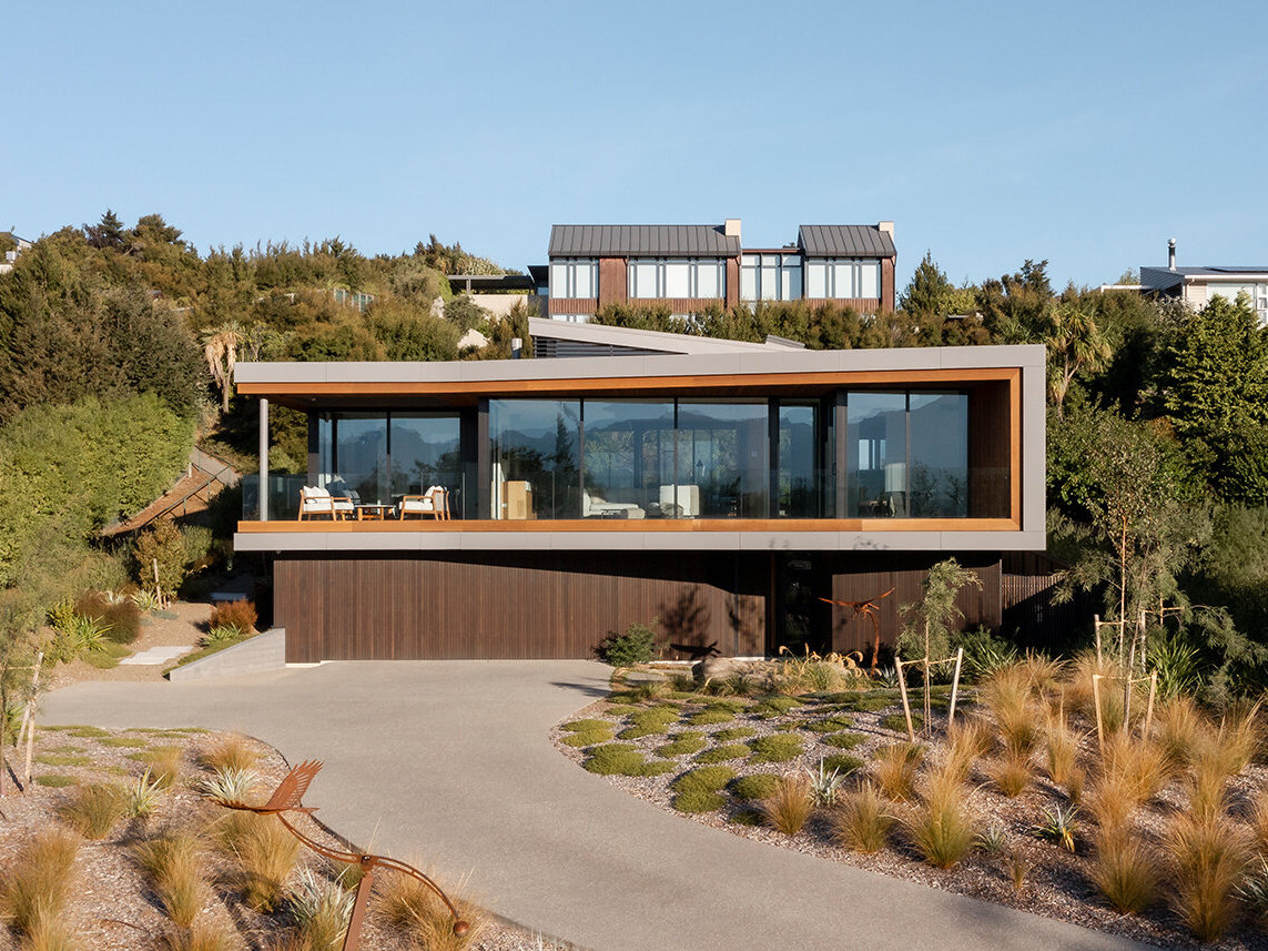 Modern two-story house featuring a light metal-framed upper floor that appears to float above a dark wood-clad base. Floor-to-ceiling windows, a covered balcony, and landscaped gardens complete the contemporary exterior.