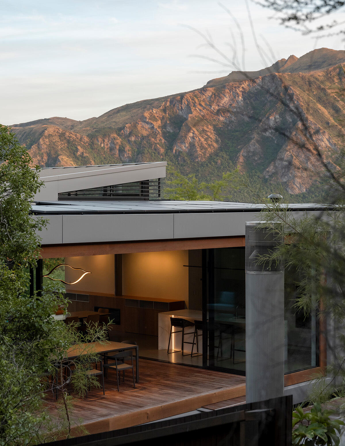 Contemporary house with an angled roof, high windows, and a covered outdoor dining area illuminated by warm interior lighting, with the mountains of W?naka rising behind it.