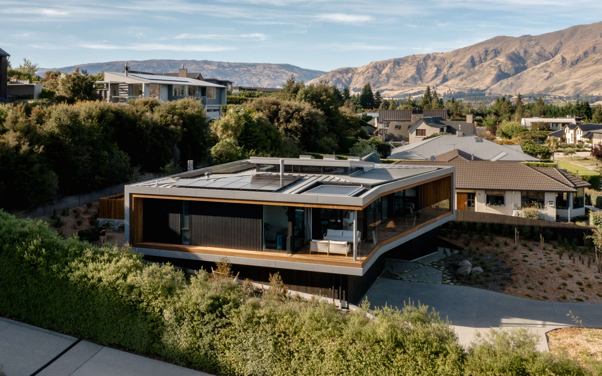 Elevated view of a contemporary W?naka house showing its angled metal roof, expansive glass walls, and sheltered outdoor living spaces. The upper floor extends above a dark-clad lower level built into the sloping property.