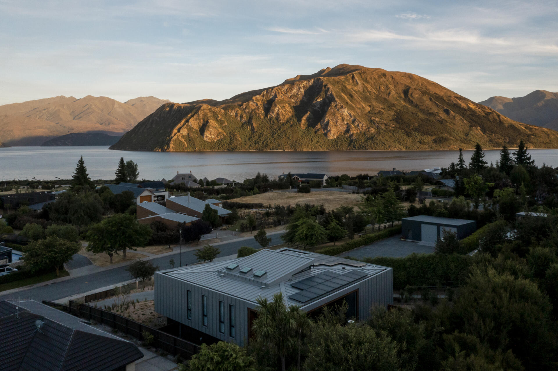 Aerial view across Lake W?naka and the surrounding mountains in New Zealand, with the metal roof of a contemporary house in the foreground and warm evening sunlight illuminating the landscape.