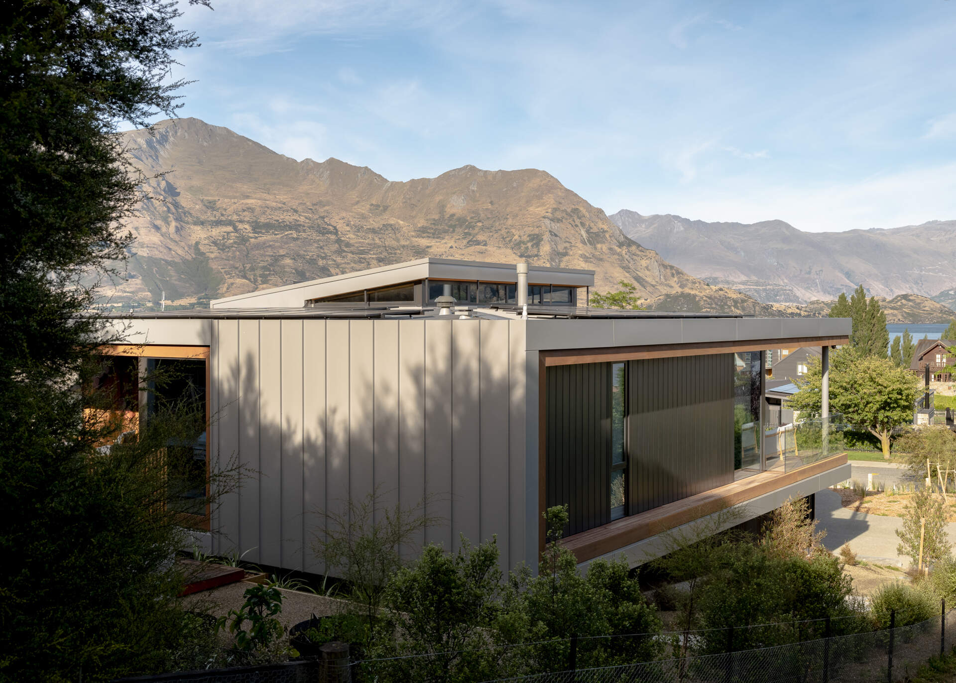 Modern W?naka house with an angled roof, light-colored metal cladding, expansive corner windows, and a covered wood balcony overlooking the surrounding landscape.
