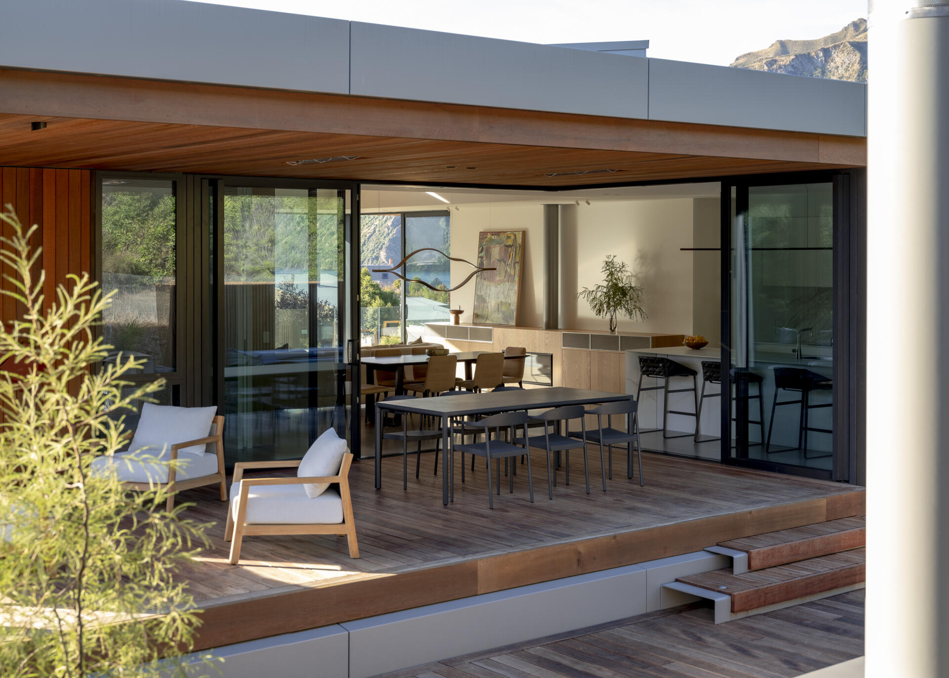 Covered outdoor living area with wood decking, contemporary lounge chairs, a black dining table, and large sliding glass doors. The sheltered terrace opens directly to the home's dining room and kitchen.