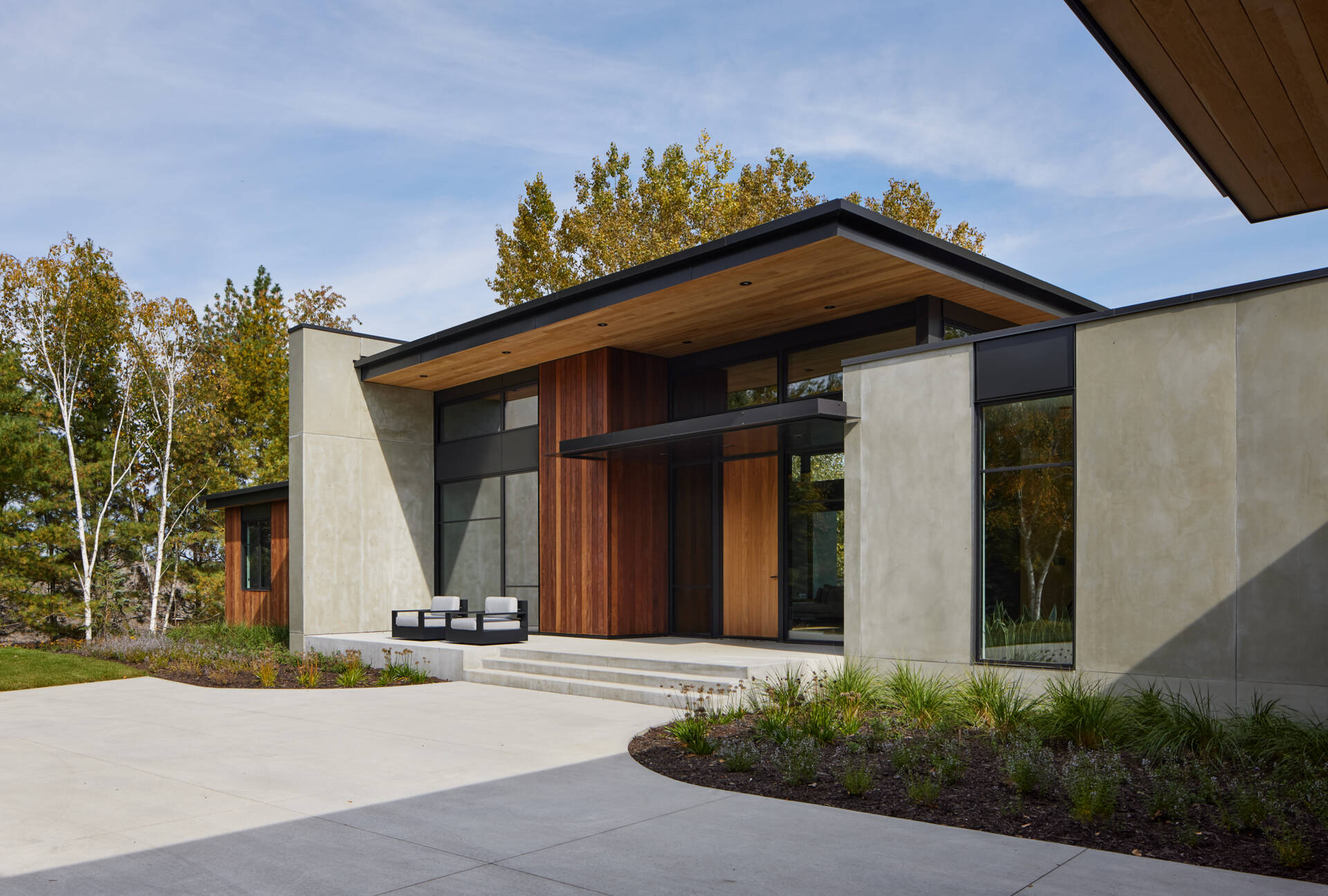 A modern house entrance featuring concrete panel walls, vertical wood siding, black-framed windows, and a flat roof with a wood-lined underside. Steps connect the covered entry to the paved courtyard.