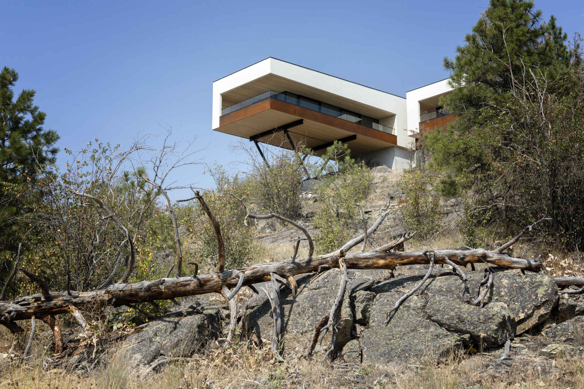 A modern cantilevered house sits above a rocky hillside in Canada's Okanagan Valley. White stucco walls, weathering steel balcony edges, glass railings, and exposed steel supports define the elevated architecture.
