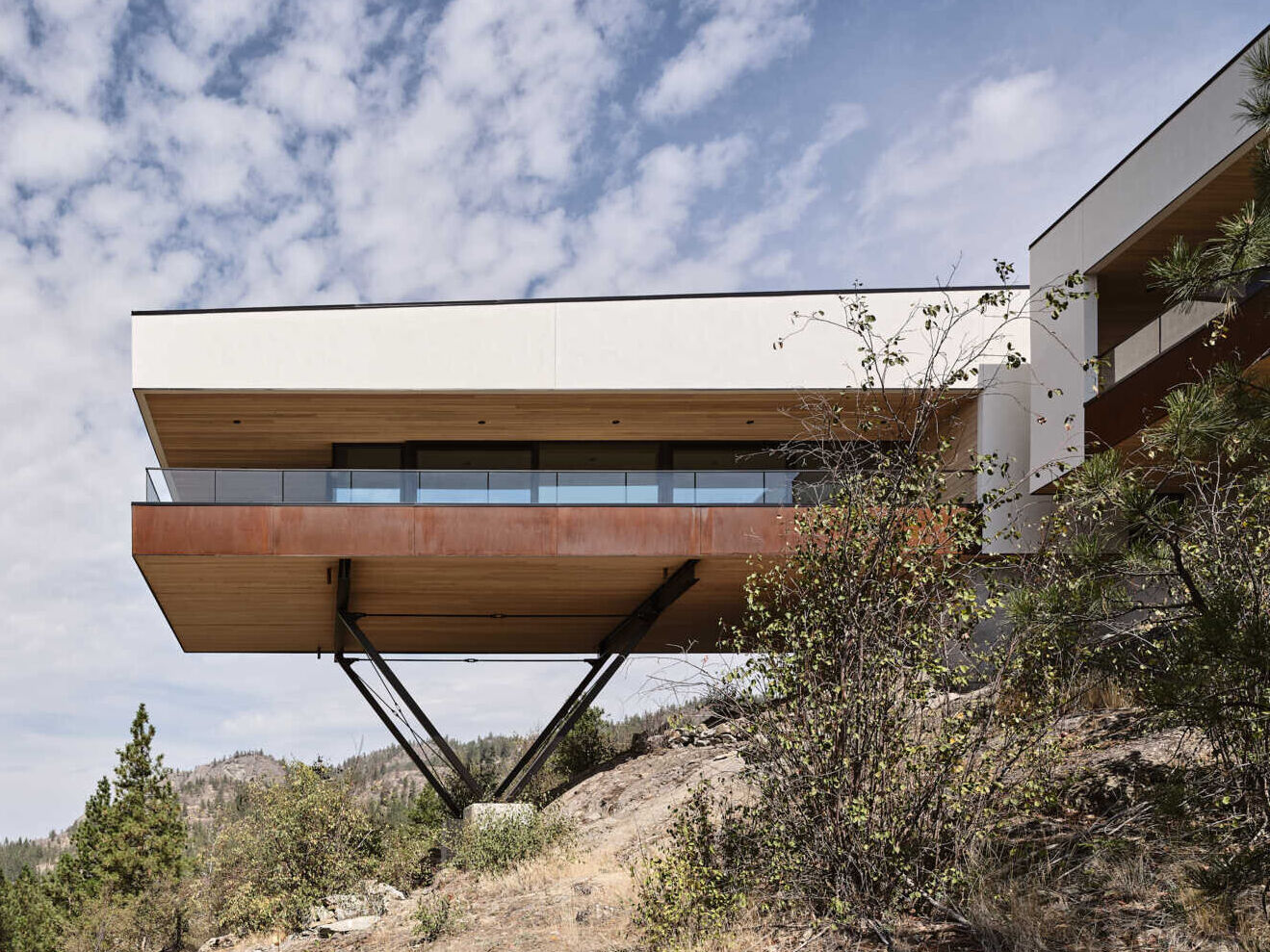 A modern cantilevered house in Naramata, Canada, extends over a steep rocky hillside on exposed V-shaped steel supports. White stucco, weathering steel, and a covered balcony define the architecture overlooking the Okanagan Valley.