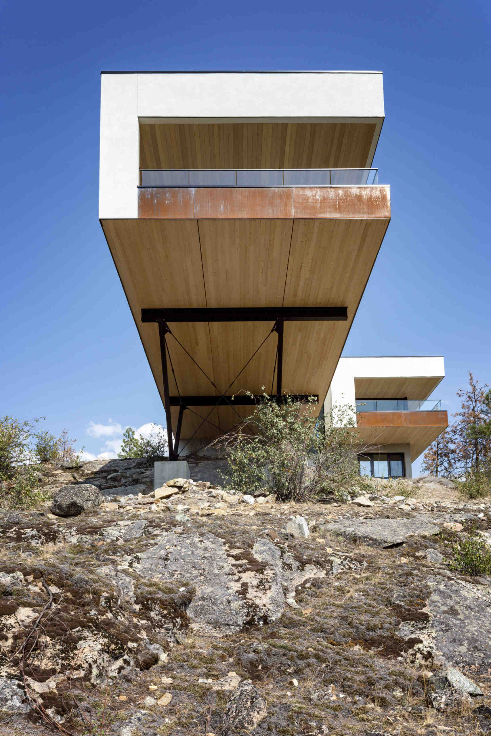 A low-angle view of a cantilevered modern house reveals its wood-lined underside and exposed steel support structure. White exterior walls, weathering steel balcony edges, and glass railings contrast with the rocky hillside.