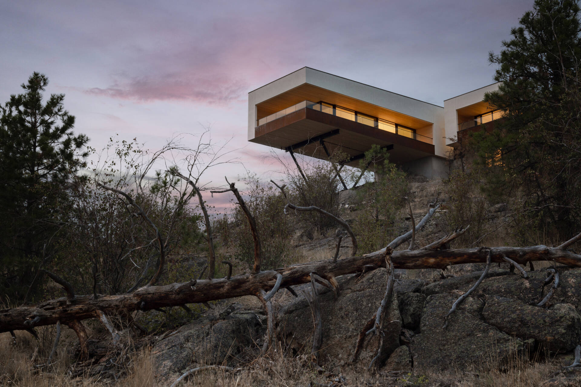 A modern cantilevered house photographed at dusk, with illuminated living spaces projecting above a rocky hillside. White exterior walls, weathering steel details, and exposed steel supports stand against a soft pink evening sky.