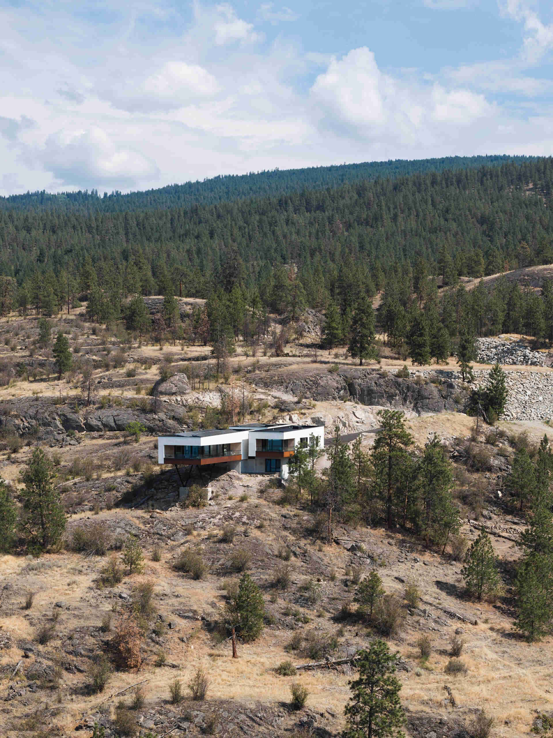 An aerial view of a modern hillside house surrounded by Ponderosa pines and rocky terrain in Canada's Okanagan Valley. The white, cantilevered building projects from the slope, with steel supports visible beneath the living wing.