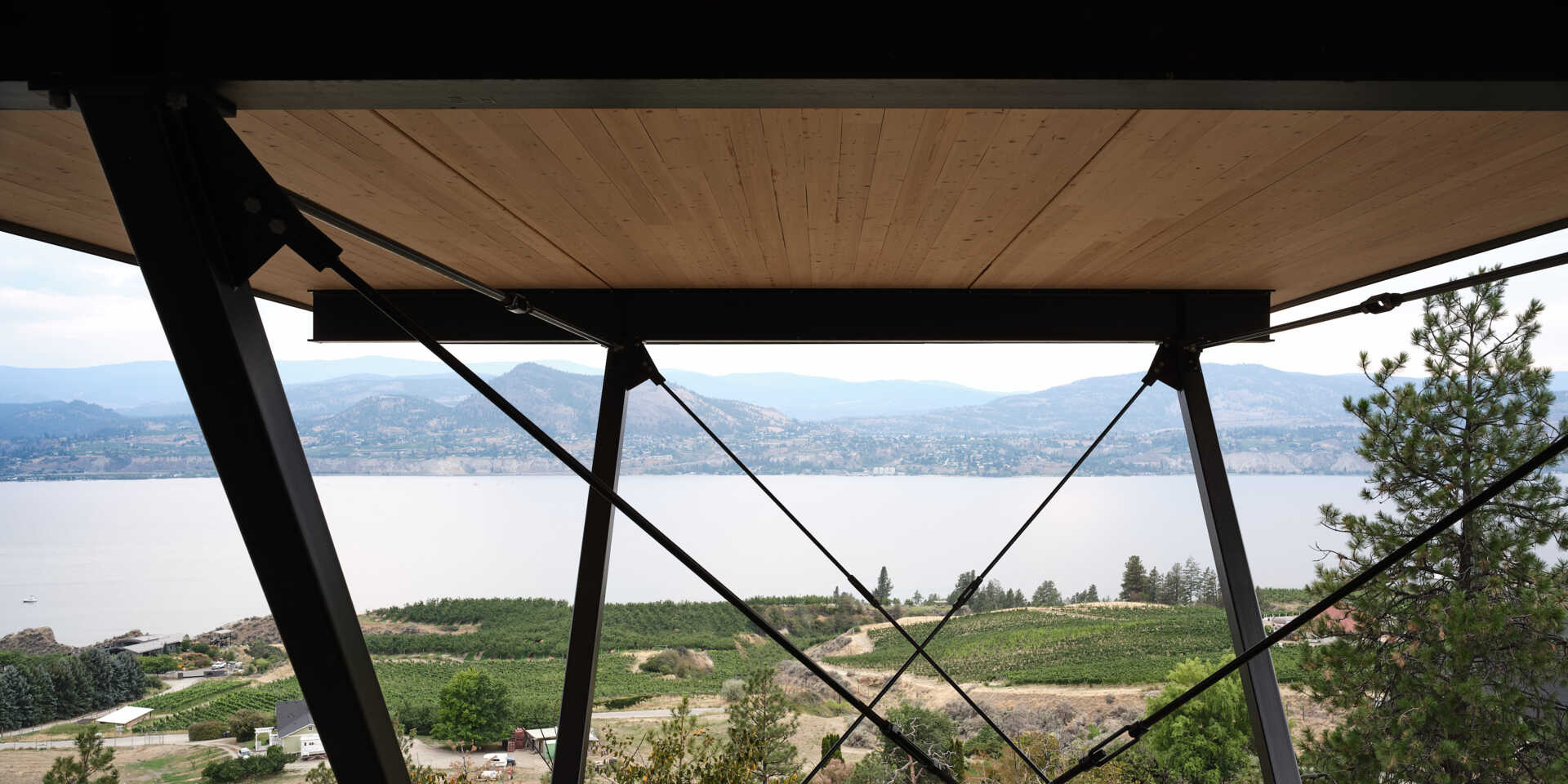 An architectural detail beneath a cantilevered house reveals black steel beams, angled braces, and tension rods supporting the elevated floor. The exposed structure frames a view across Okanagan Lake and the surrounding mountains.