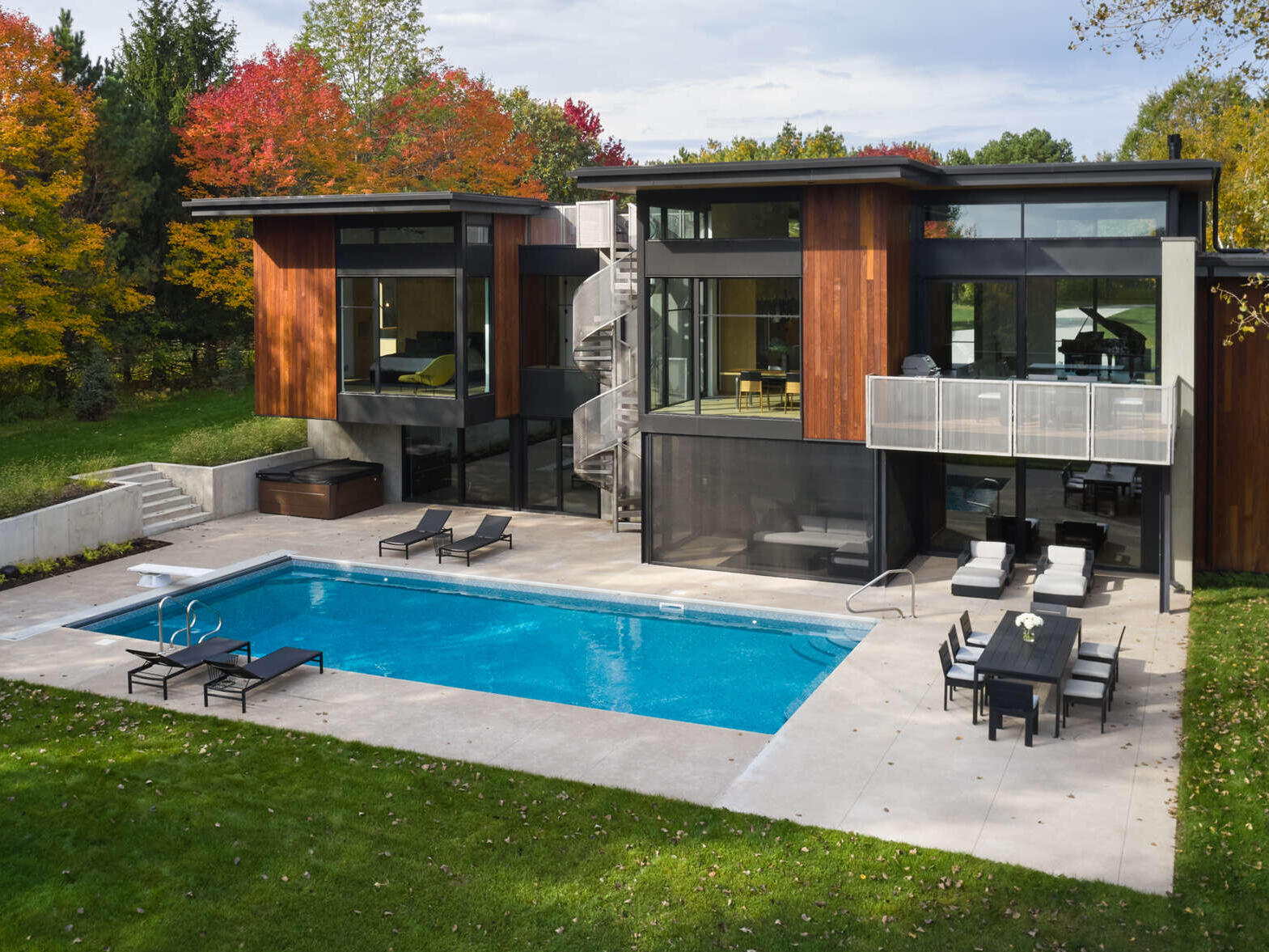 A modern Minnesota house with flat roofs, wood siding, large windows, and a stainless steel spiral staircase. A rectangular swimming pool and concrete patio sit beside the home, surrounded by colorful autumn trees.