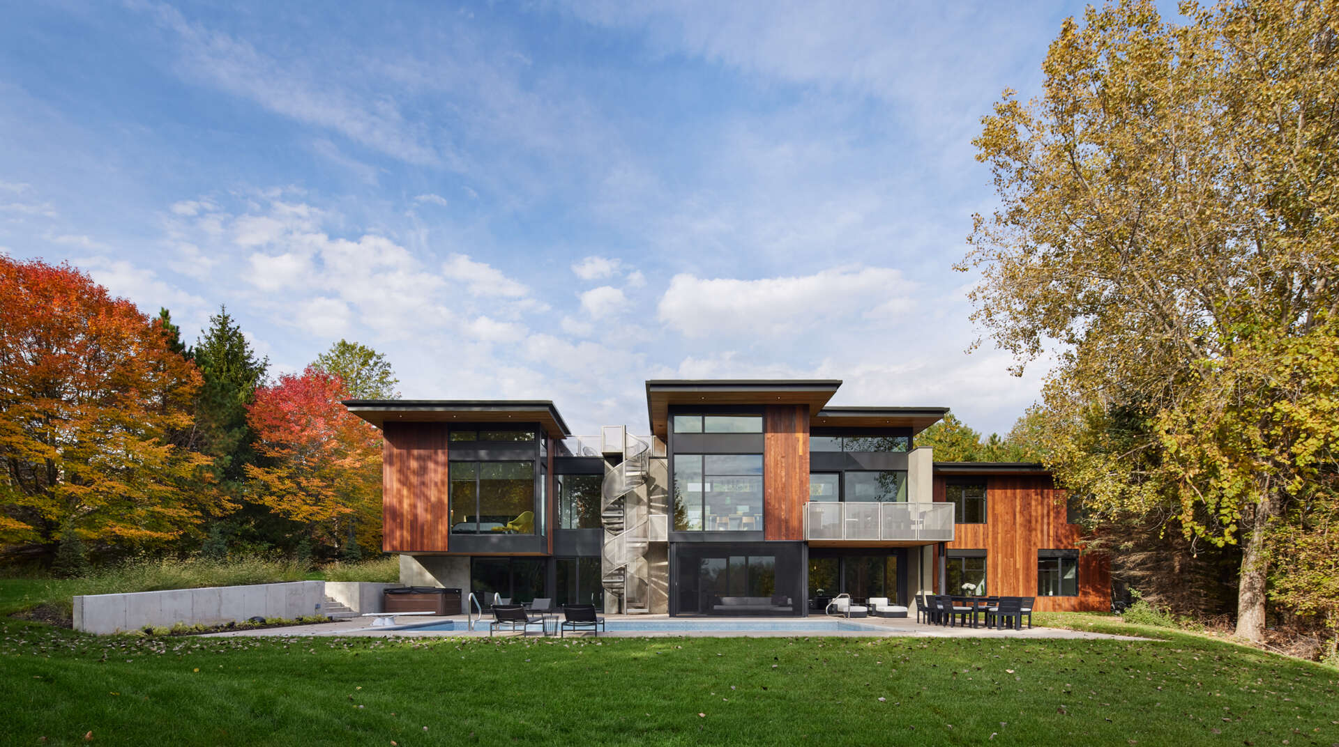 A modern house with a swimming pool, flat roofs, large windows, wood siding, and a central exterior spiral staircase. Outdoor seating areas and a balcony overlook the concrete pool patio.