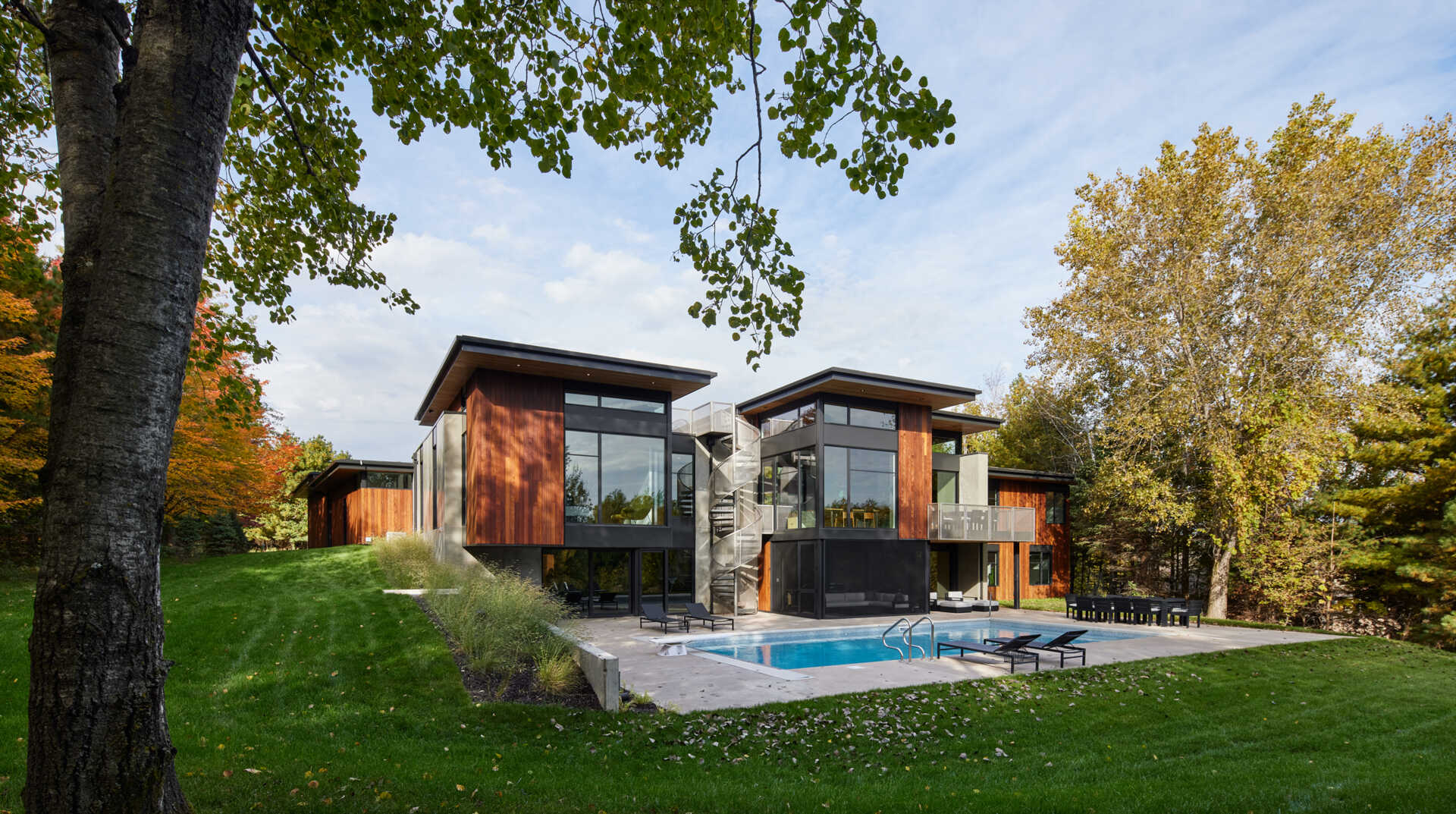 A contemporary house surrounded by mature trees, featuring projecting upper-level rooms, wood siding, large glass walls, and an exterior spiral staircase. A rectangular swimming pool extends across the rear patio.