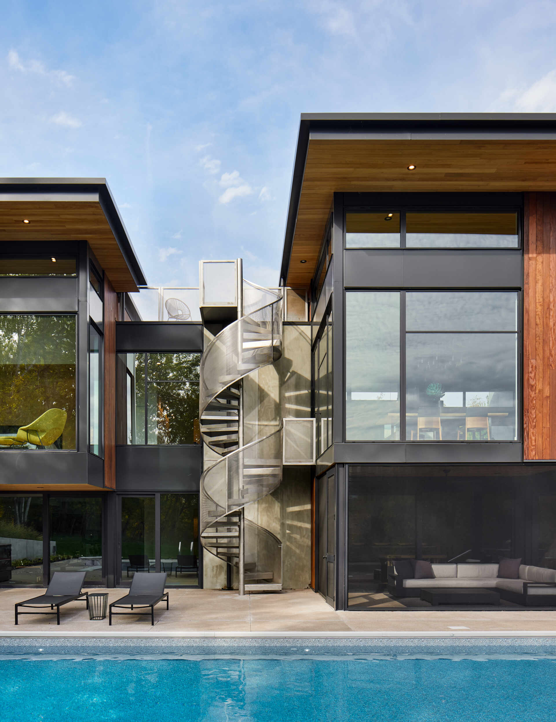 A stainless steel outdoor spiral staircase rises between two sections of a contemporary house. Flat roof overhangs, large black-framed windows, and a swimming pool surround the sculptural metal stairway.