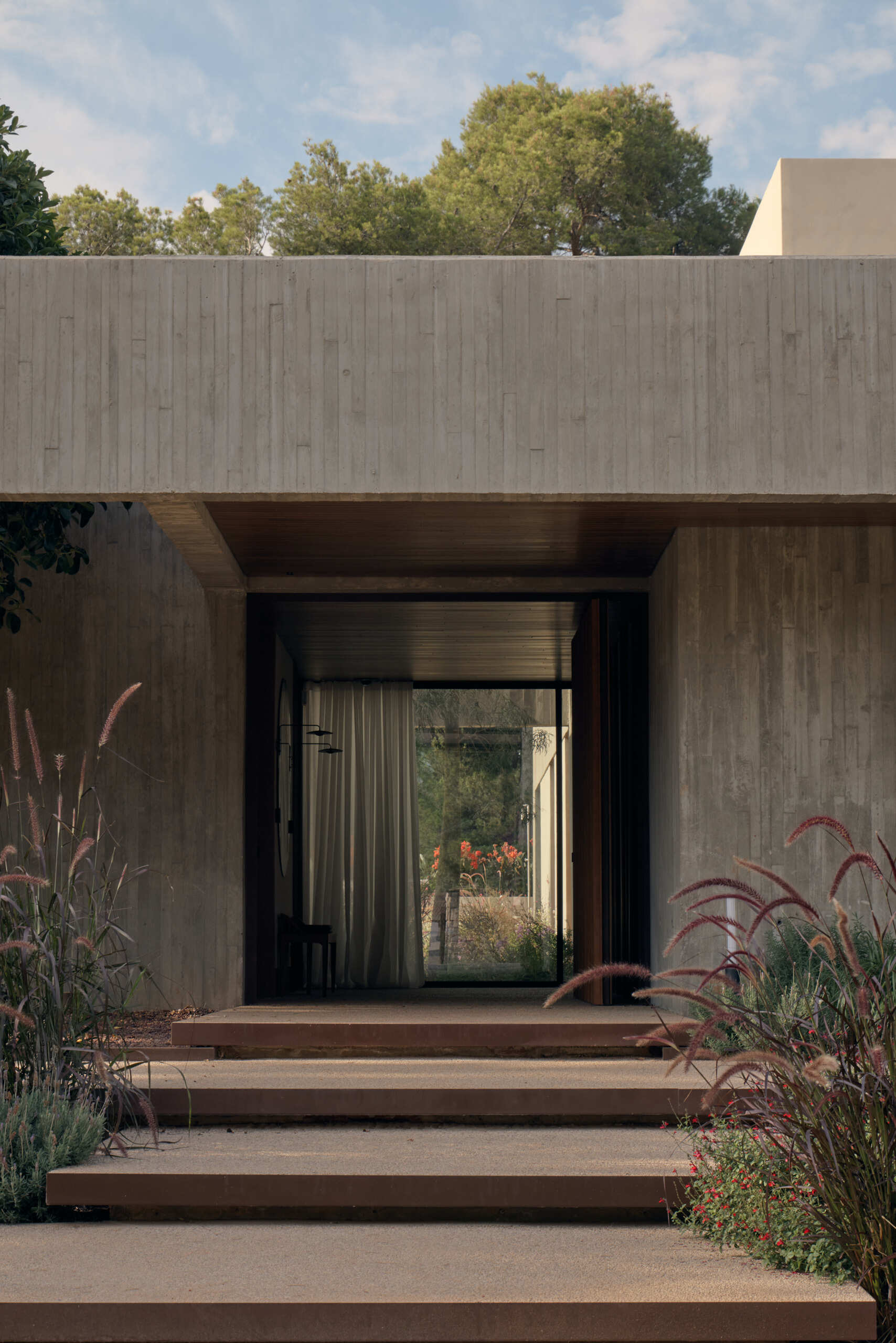 Step inside Casa Tossal and explore its modern entryway, featuring floating-style concrete garden steps, wood ceilings, large wooden doors, sculptural artwork, and a minimalist wooden bench.