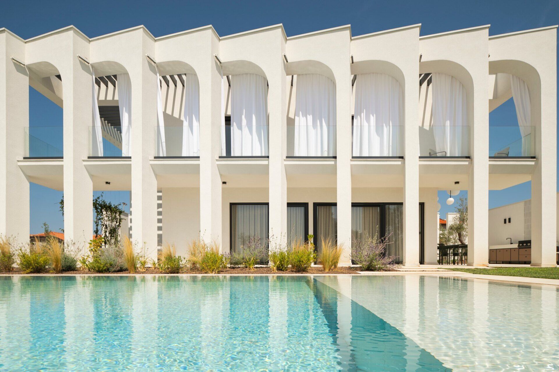Explore the swimming pool and south-facing façade of Arc House, a modern villa in Istria, Croatia. A rectangular pool, sculptural concrete arches, covered balconies and Mediterranean landscaping create an impressive contemporary outdoor living area.
