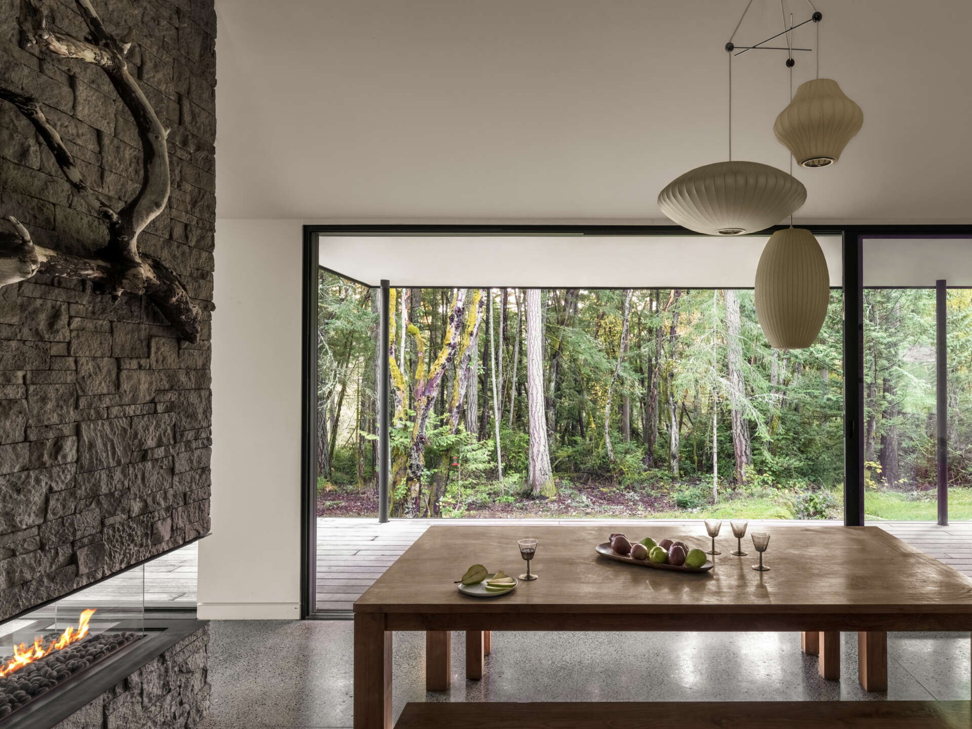 A modern dining area with a large wooden table, bench seating, sculptural pendant lights, polished concrete floors, and sliding glass doors overlooking a Pacific Northwest forest.