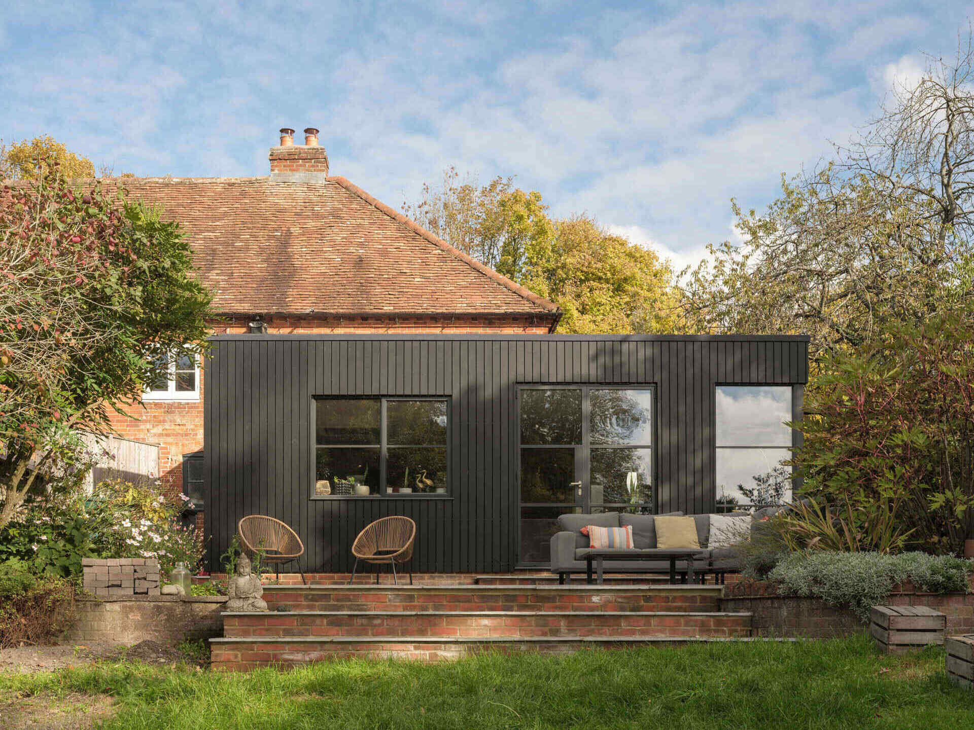 A 200-year-old cottage in Hampshire features a modern black-clad extension with vertical siding, a flat roof, corner glazing, and an outdoor terrace overlooking the garden.