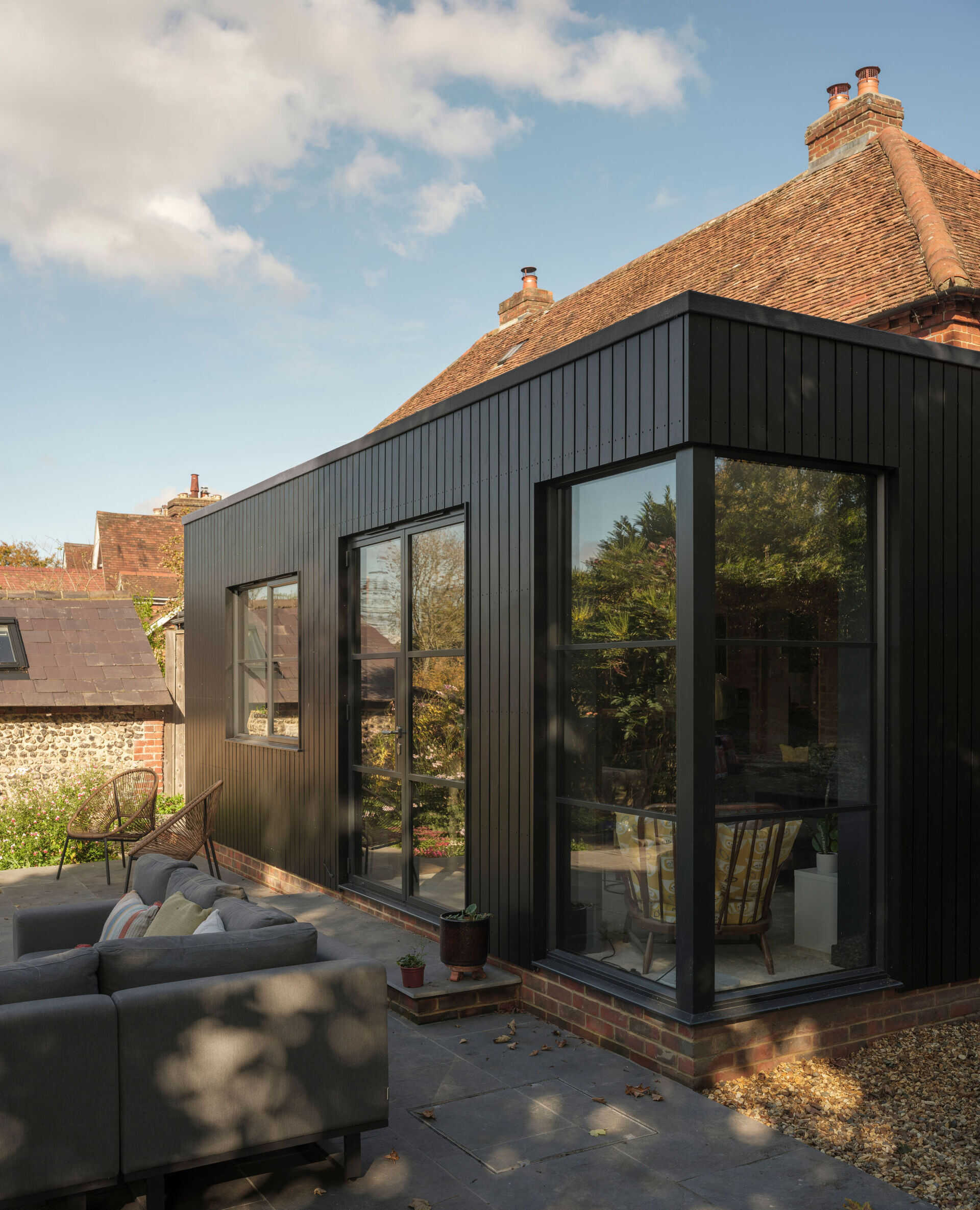 A 200-year-old cottage in Hampshire features a modern black-clad extension with vertical siding, a flat roof, corner glazing, and an outdoor terrace overlooking the garden.