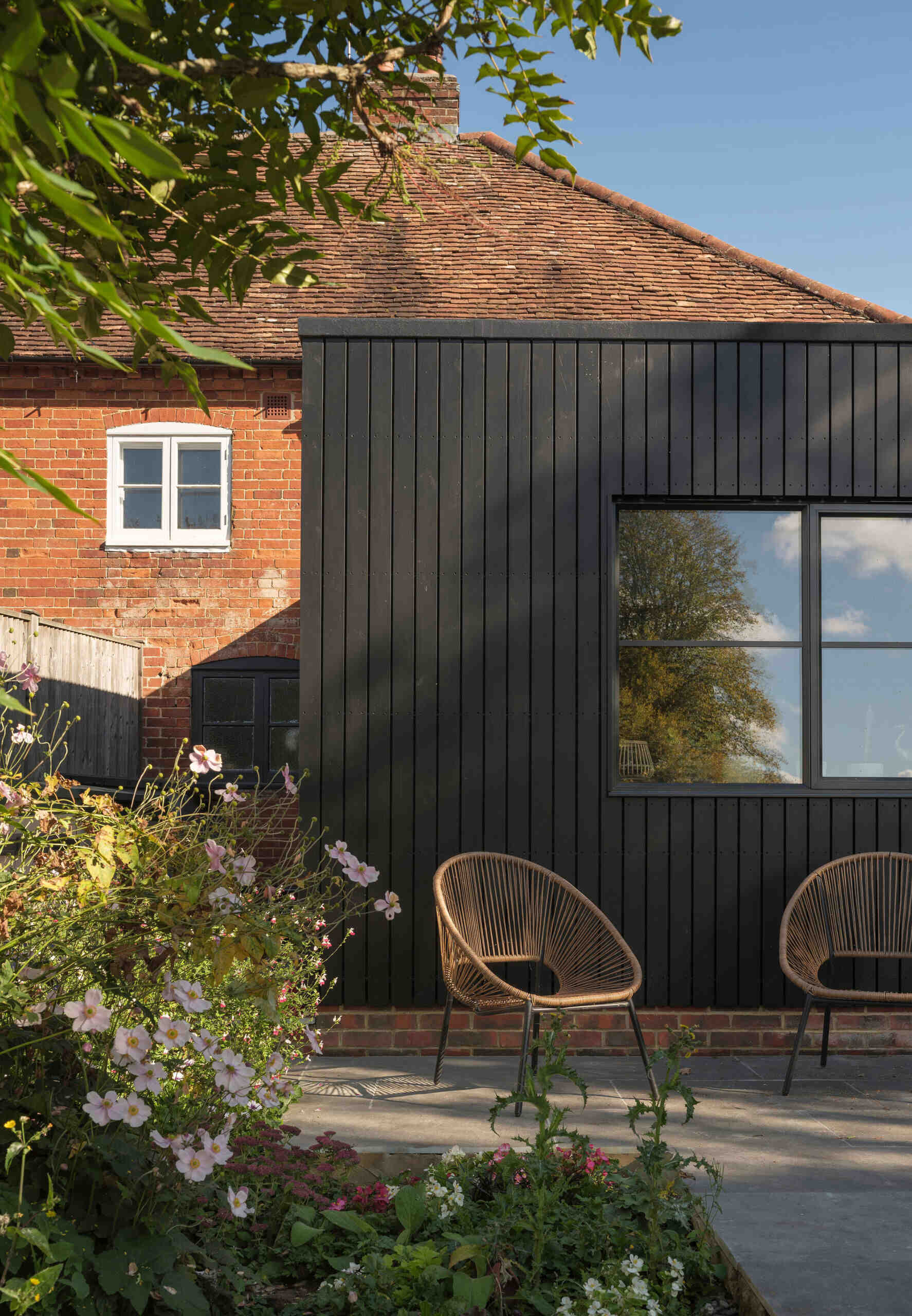 A 200-year-old cottage in Hampshire features a modern black-clad extension with vertical siding, a flat roof, corner glazing, and an outdoor terrace overlooking the garden.