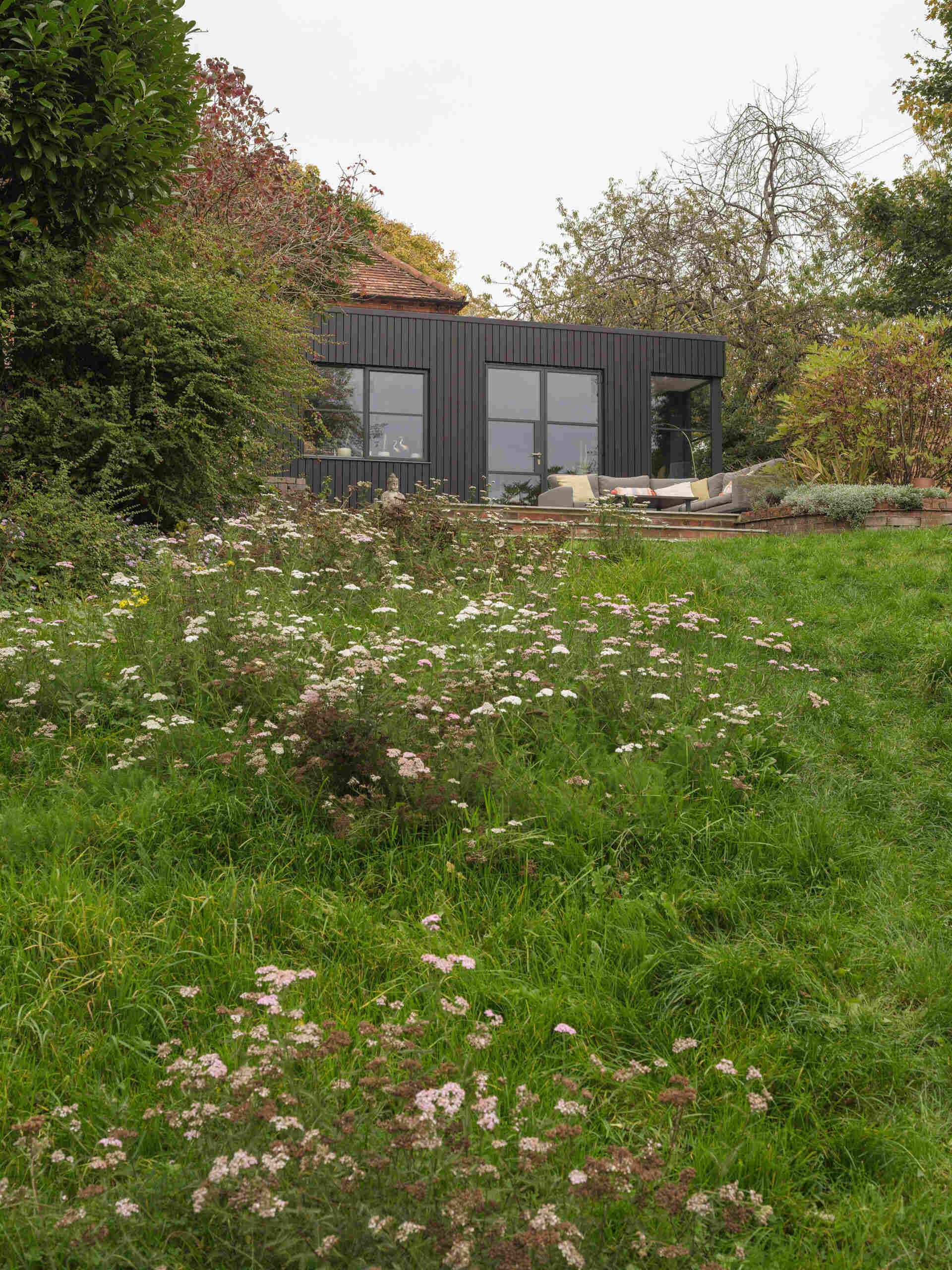 A 200-year-old cottage in Hampshire features a modern black-clad extension with vertical siding, a flat roof, corner glazing, and an outdoor terrace overlooking the garden.