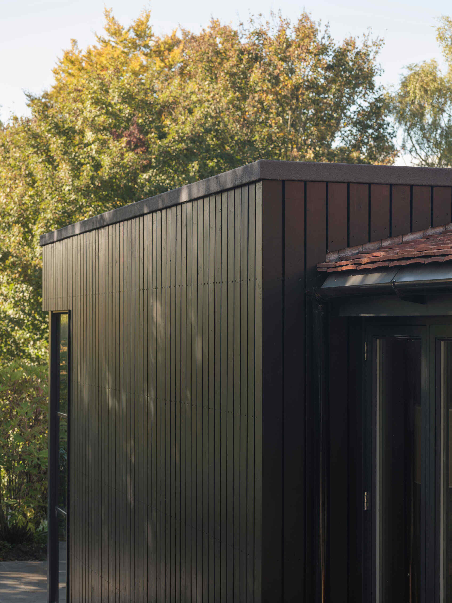 A 200-year-old cottage in Hampshire features a modern black-clad extension with vertical siding, a flat roof, corner glazing, and an outdoor terrace overlooking the garden.