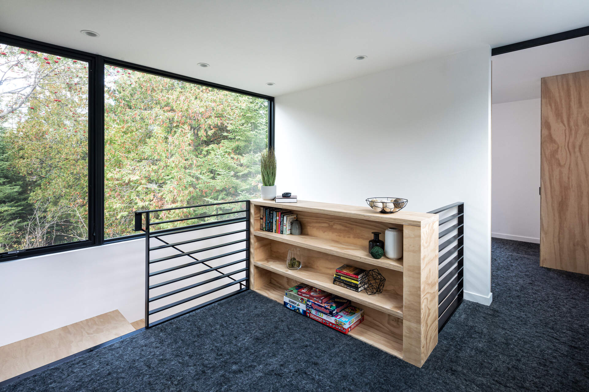 A small upstairs reading area with a plywood bookshelf, black metal stair railing, gray carpet tiles, and a large window overlooking the surrounding trees. A sliding plywood door leads toward the bedrooms.