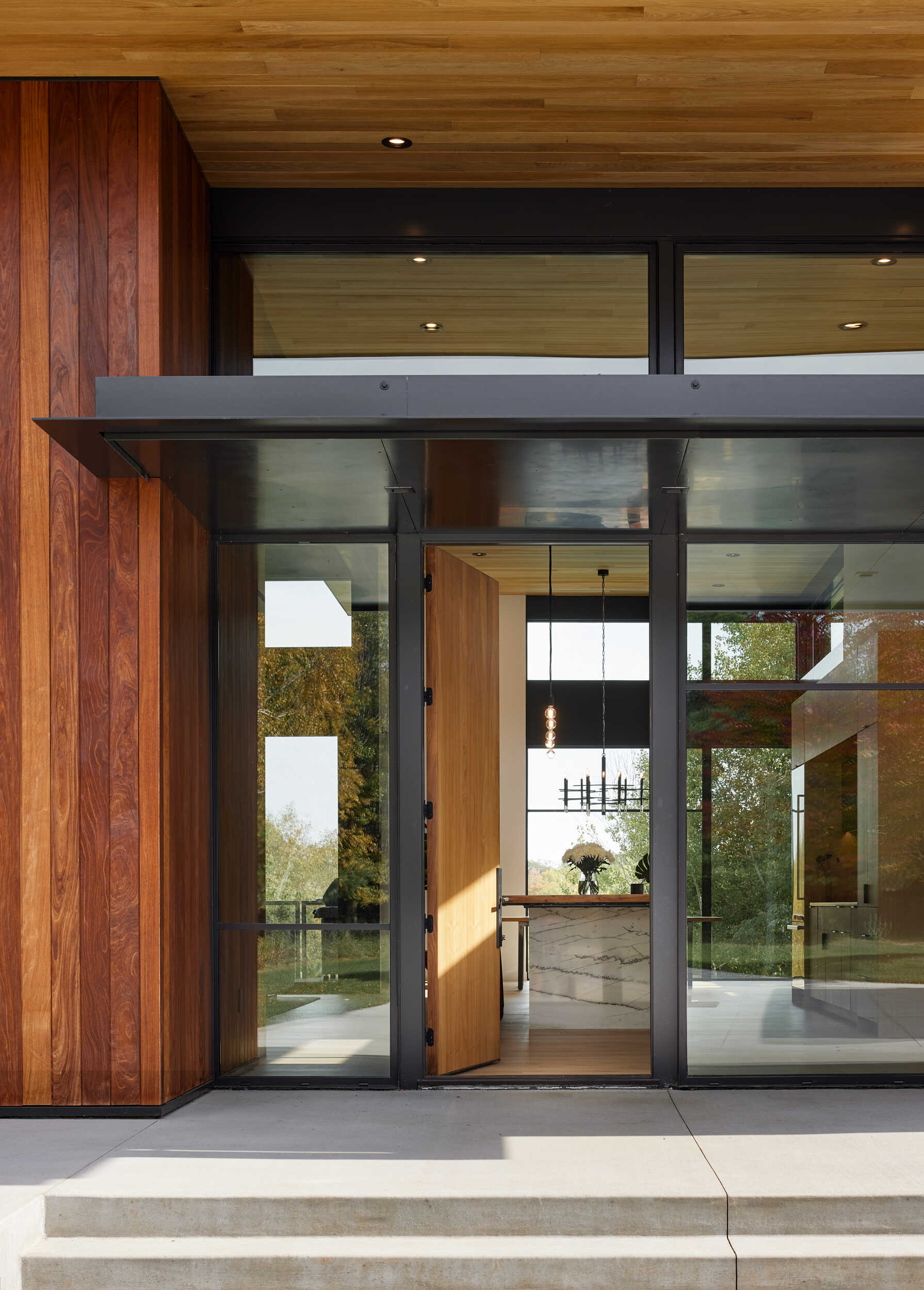 A contemporary wood front door set within a wall of black-framed glass, with a dark metal canopy above. The entrance reveals views through to the home's modern kitchen and dining area.
